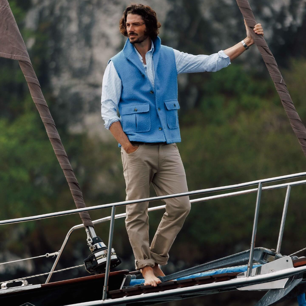 A man with wavy hair stands barefoot on a sailboat, wearing beige pants, a white shirt, and a light blue Campbells of Beauly vest, with one hand resting on a sail and a scenic, blurred background of rocks and greenery.