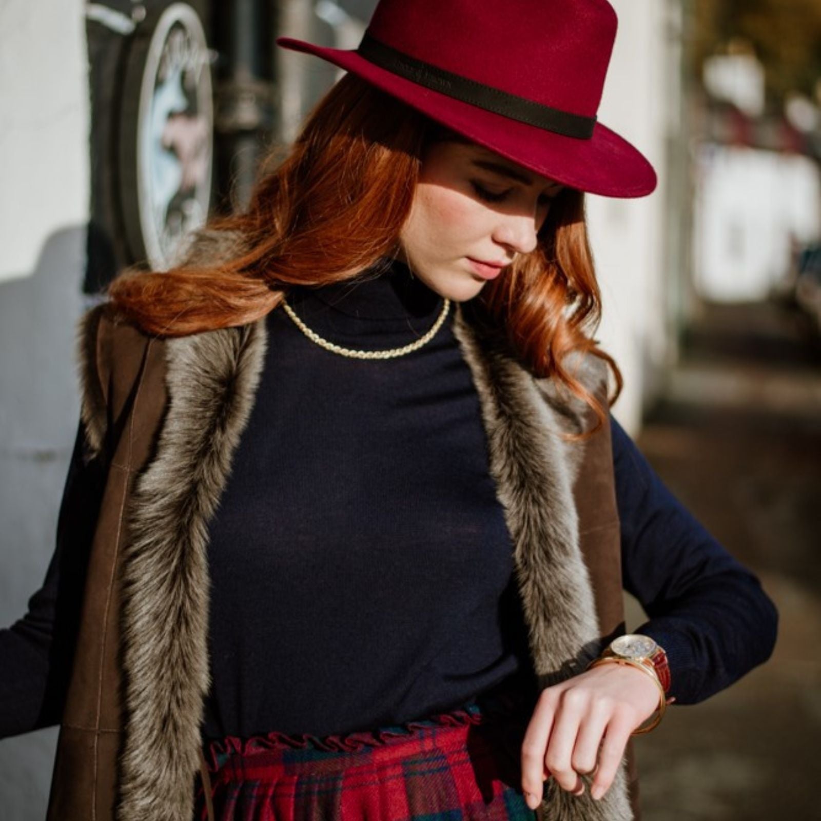 A woman with long red hair wears a burgundy hat, a fur-trimmed vest, a navy turtleneck, and a red plaid Campbells of Beauly skirt. She looks down at her wristwatch while standing outdoors in sunlight.
