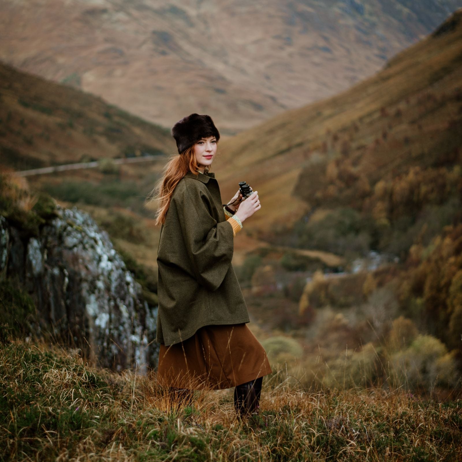 A woman in a Campbells of Beauly olive green coat and brown hat stands on a grassy hillside holding binoculars, with rolling hills and a winding road in the background on an overcast day.