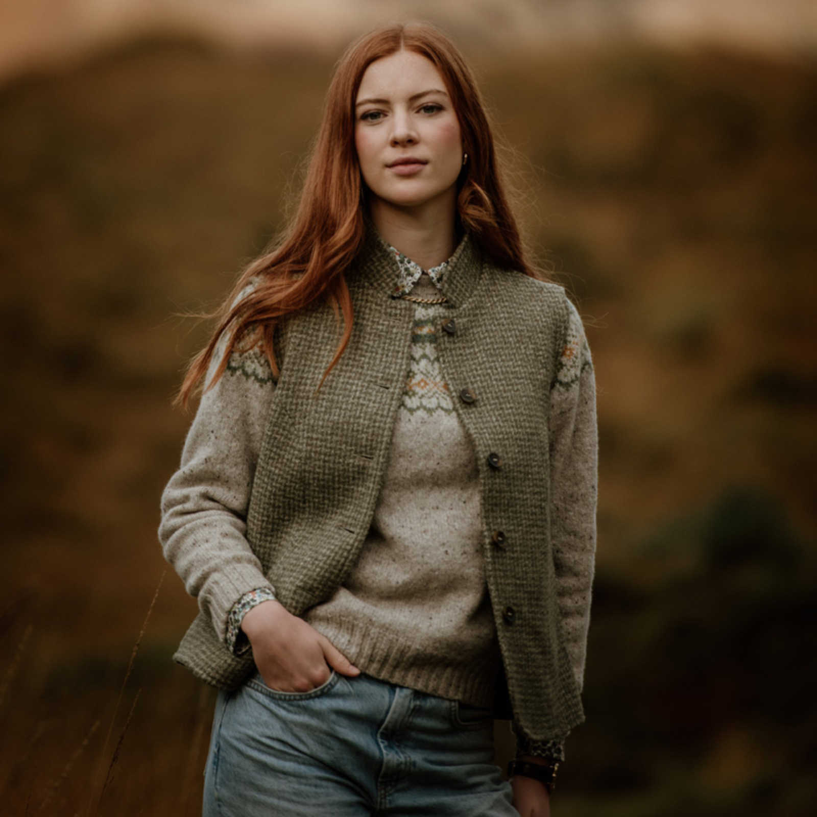 A young woman with long red hair stands outdoors, wearing a patterned sweater, buttoned vest from Campbells of Beauly, and jeans. The background is softly blurred, showing earthy tones and natural scenery.