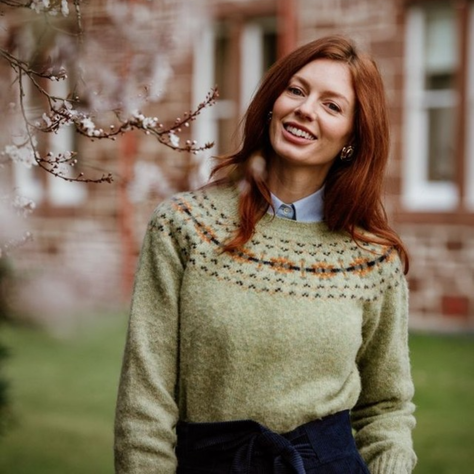 A woman with long red hair smiles outdoors, wearing a light green patterned sweater over a collared shirt. Blurred brick building and blooming tree branches are visible in the background.