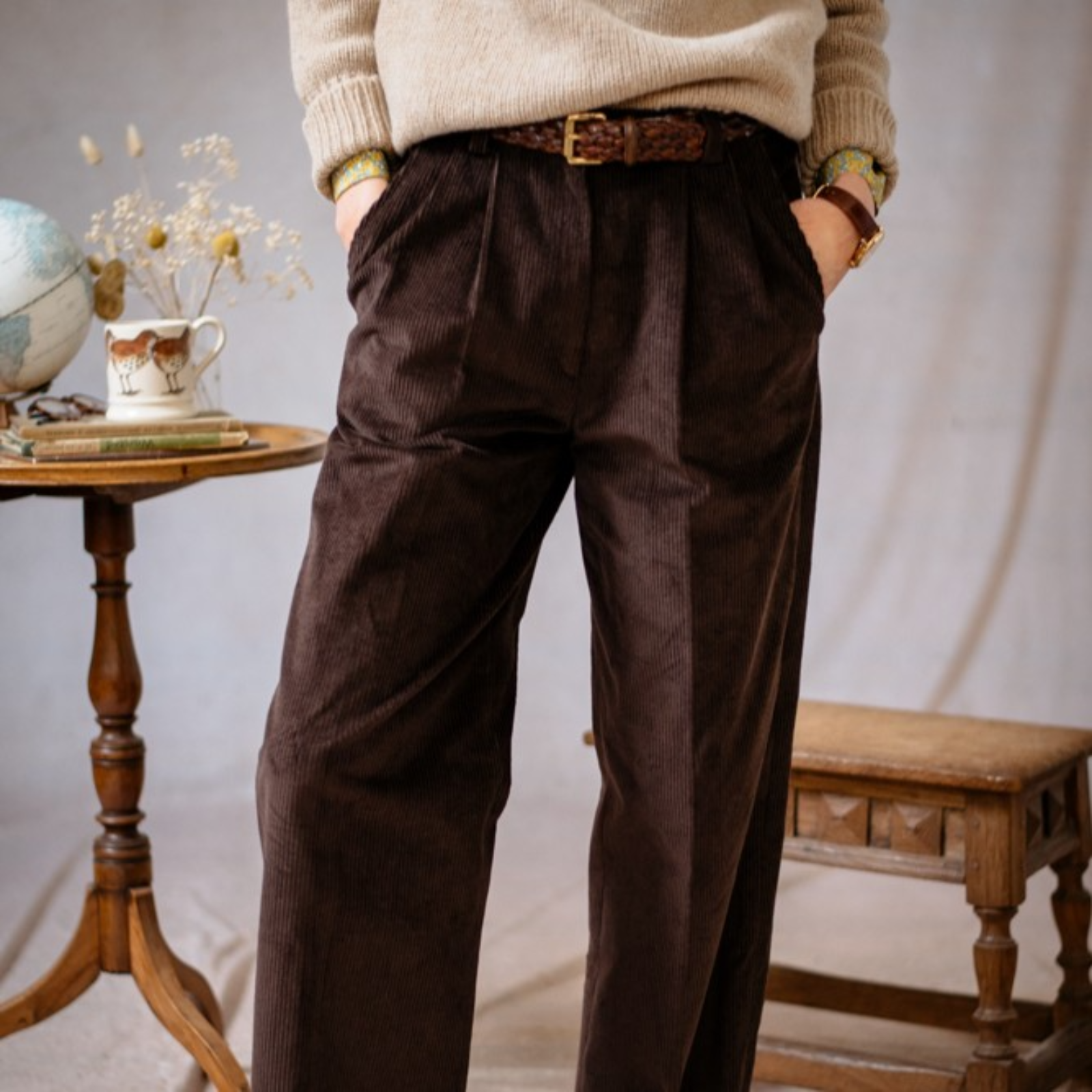 A person wearing a beige sweater and high-waisted brown corduroy trousers stands with hands in pockets beside a wooden table holding a teacup, saucer, globe, and vase, with a wooden stool nearby.