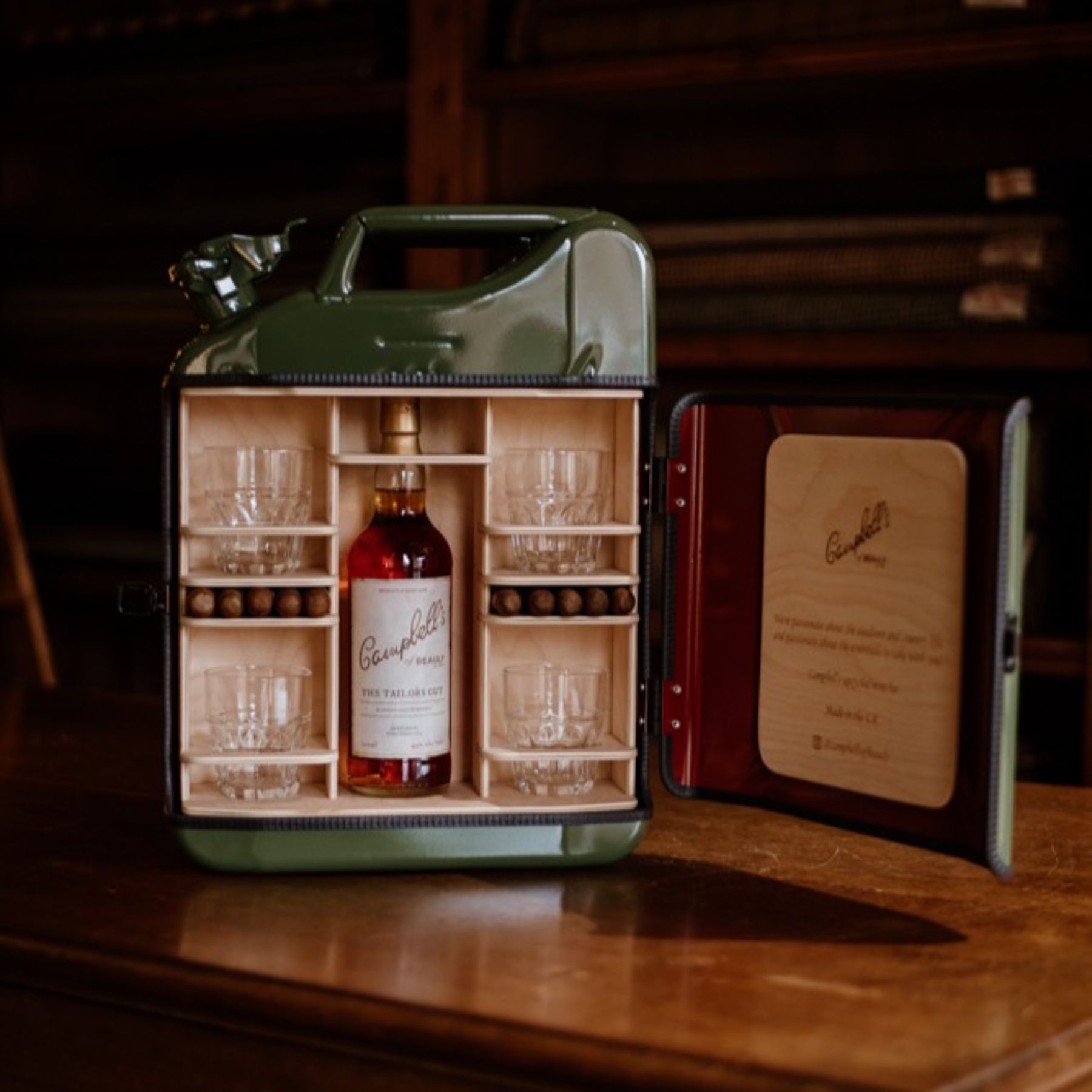 A green metal jerry can repurposed as a portable bar, open to reveal a bottle of Campbell’s whisky, four glasses, and compartments with brown stones, displayed on a wooden table with shelves in the background.