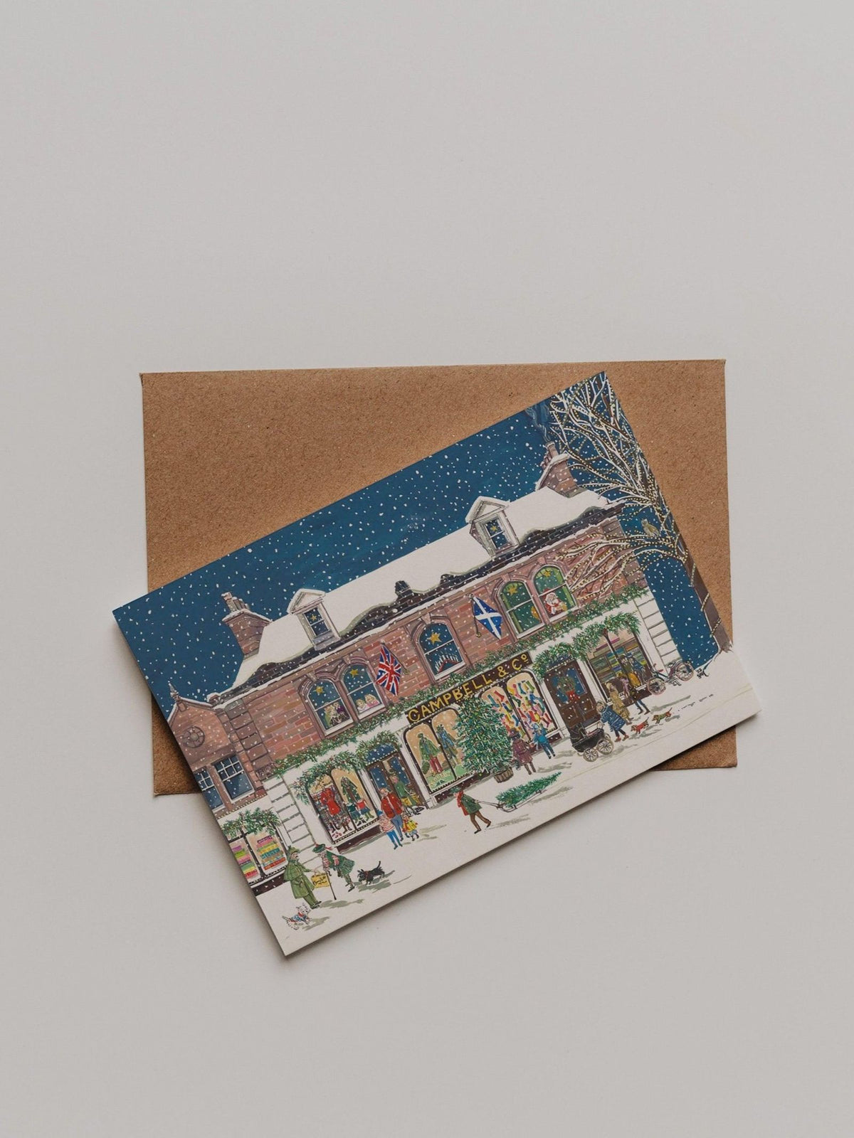 The Campbell’s at Christmas Greeting Card from Campbell’s of Beauly rests atop a brown envelope on a light surface, showcasing a snowy store scene and festive flags. The elegant illustration by Sophie Murray enhances the charm of this seasonal thank you card.
