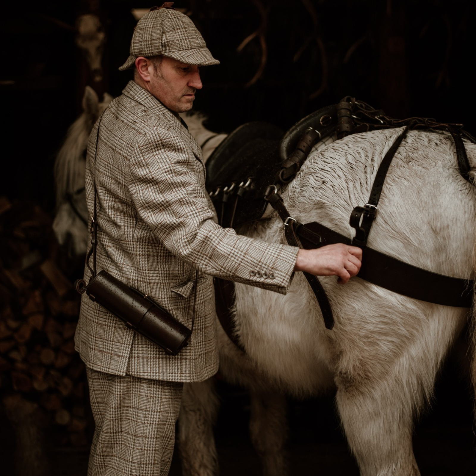 A man in a plaid suit and cap from Campbells of Beauly adjusts the harness on a white horse. He carries a cylindrical case over his shoulder, with stacked logs in the background.