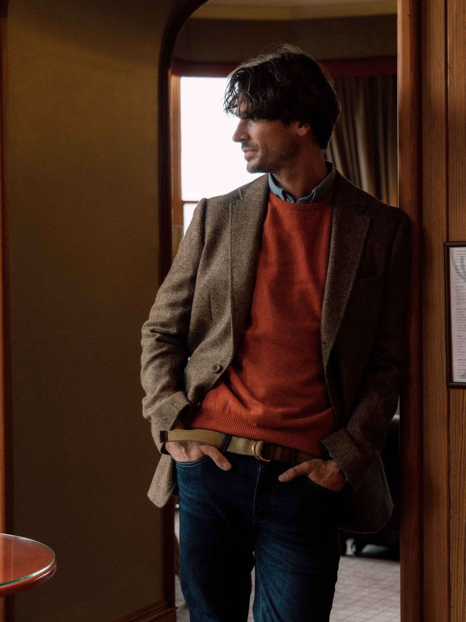 A man with wavy hair stands indoors by a curtained window, wearing a brown blazer over a Campbell's of Beauly Lambswool Crew Neck Jumper in orange, layered above a blue shirt, paired with blue jeans and a tan belt, hands in his pockets and looking to the side.