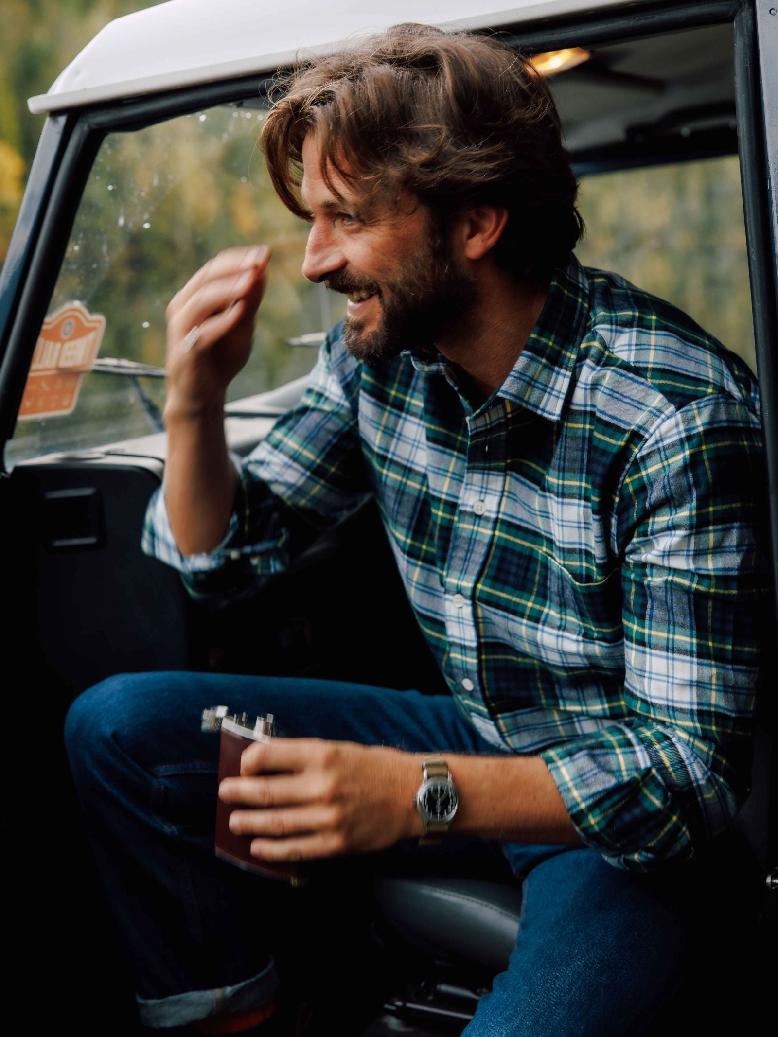 A smiling man with wavy brown hair and a beard wears a Campbell's of Beauly Tartan Shirt and jeans, sitting in a vehicle doorway and holding a flask.
