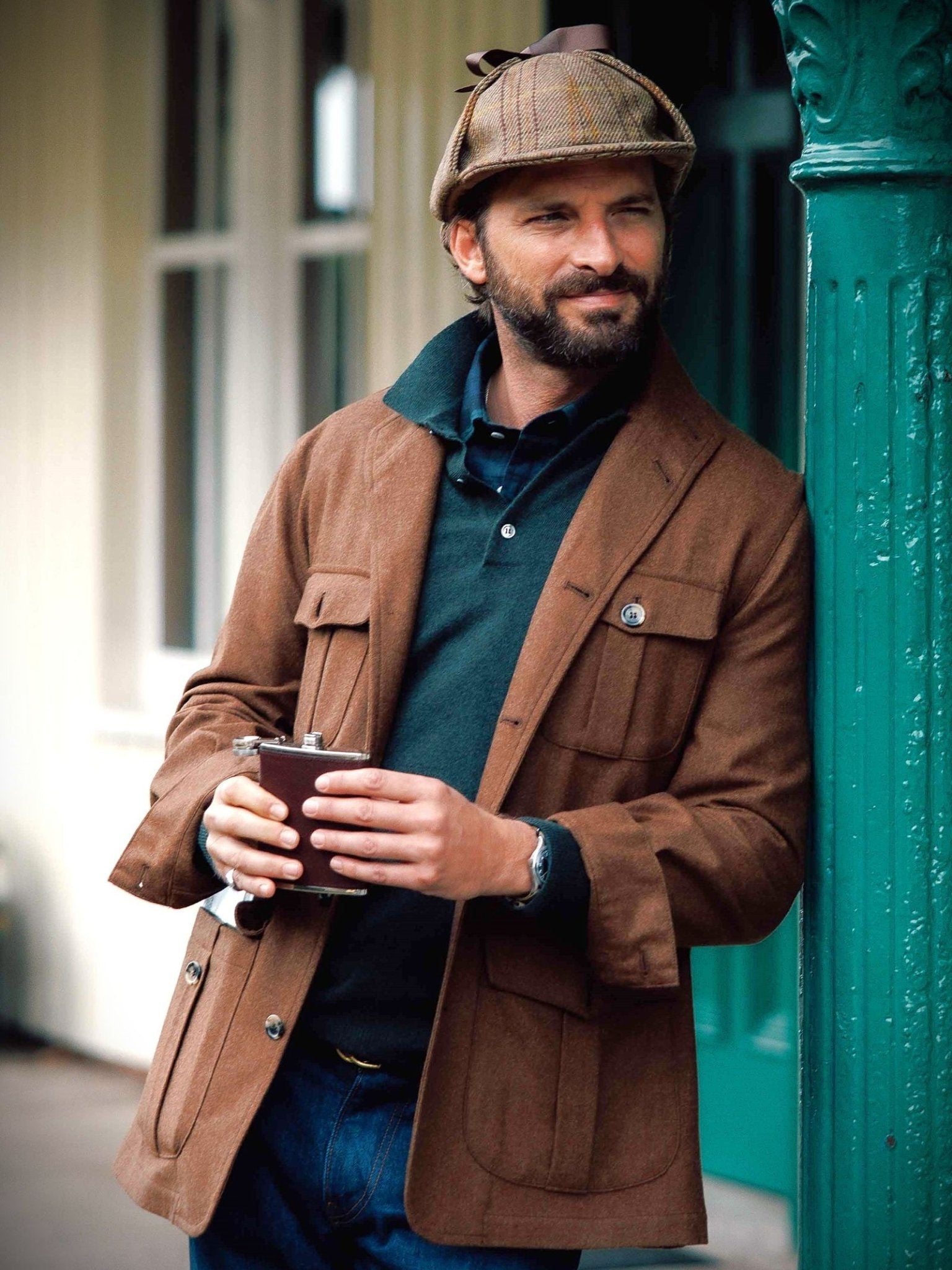 A bearded man in a brown jacket, tweed cap, and Campbell's of Beauly Merino & Cashmere Shirt Jumper leans against a green post, holding a silver flask and smiling slightly.