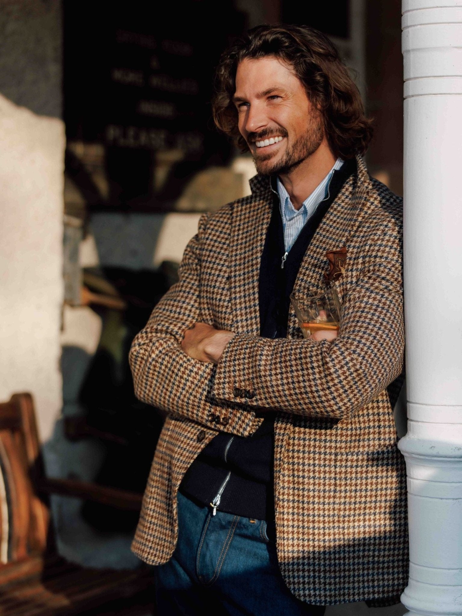 A smiling man with wavy hair stands arms crossed, wearing a Campbells of Beauly Navy Gun Club Tweed Jacket over a blue shirt and jeans, leaning casually against a white column outdoors.