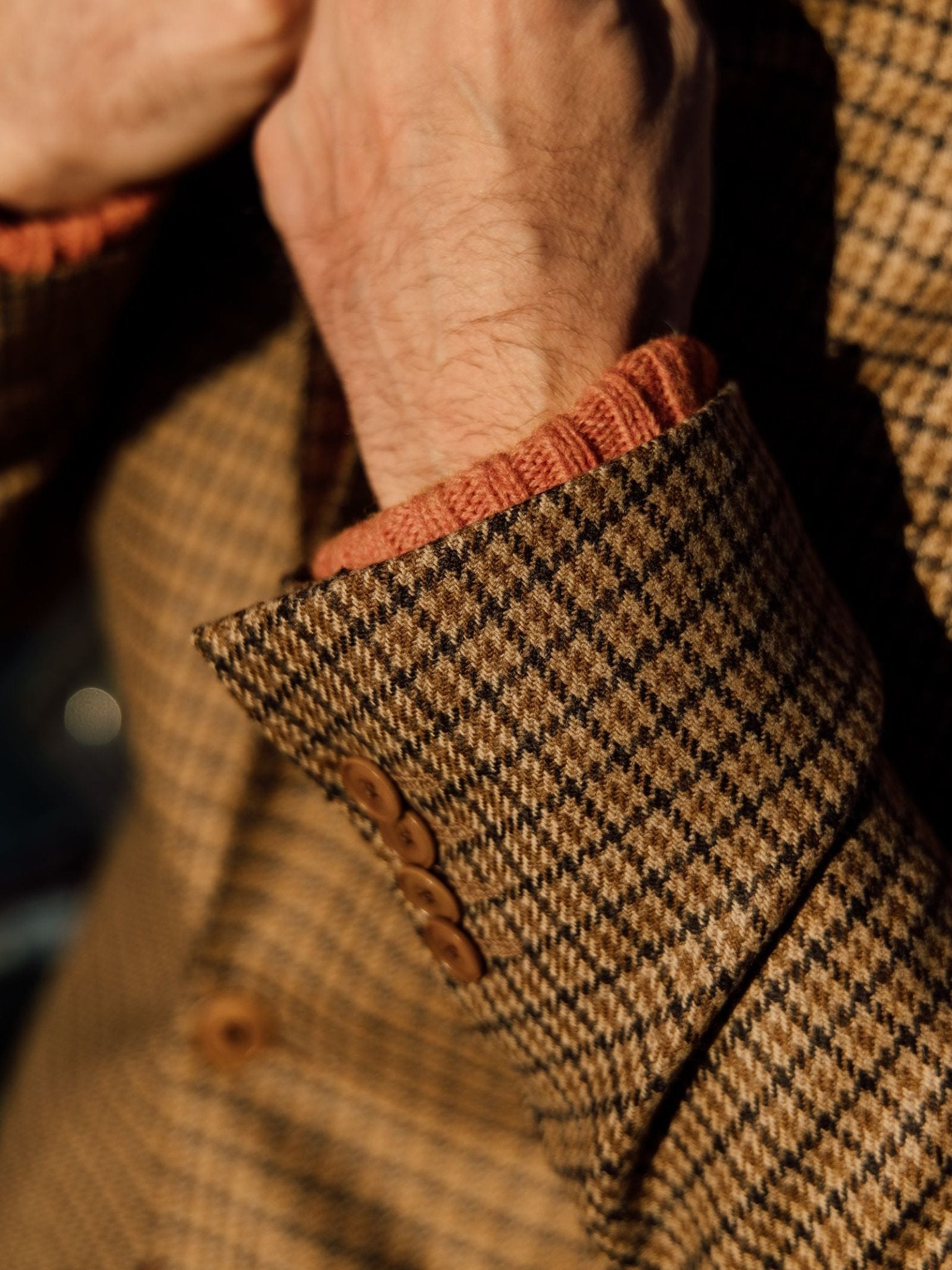 A close-up shows a person wearing a Campbells of Beauly Gun Club Jacket in brown houndstooth, regular fit, with brown buttons over a rust knit sweater. Their hands are partly visible as they adjust the jacket.
