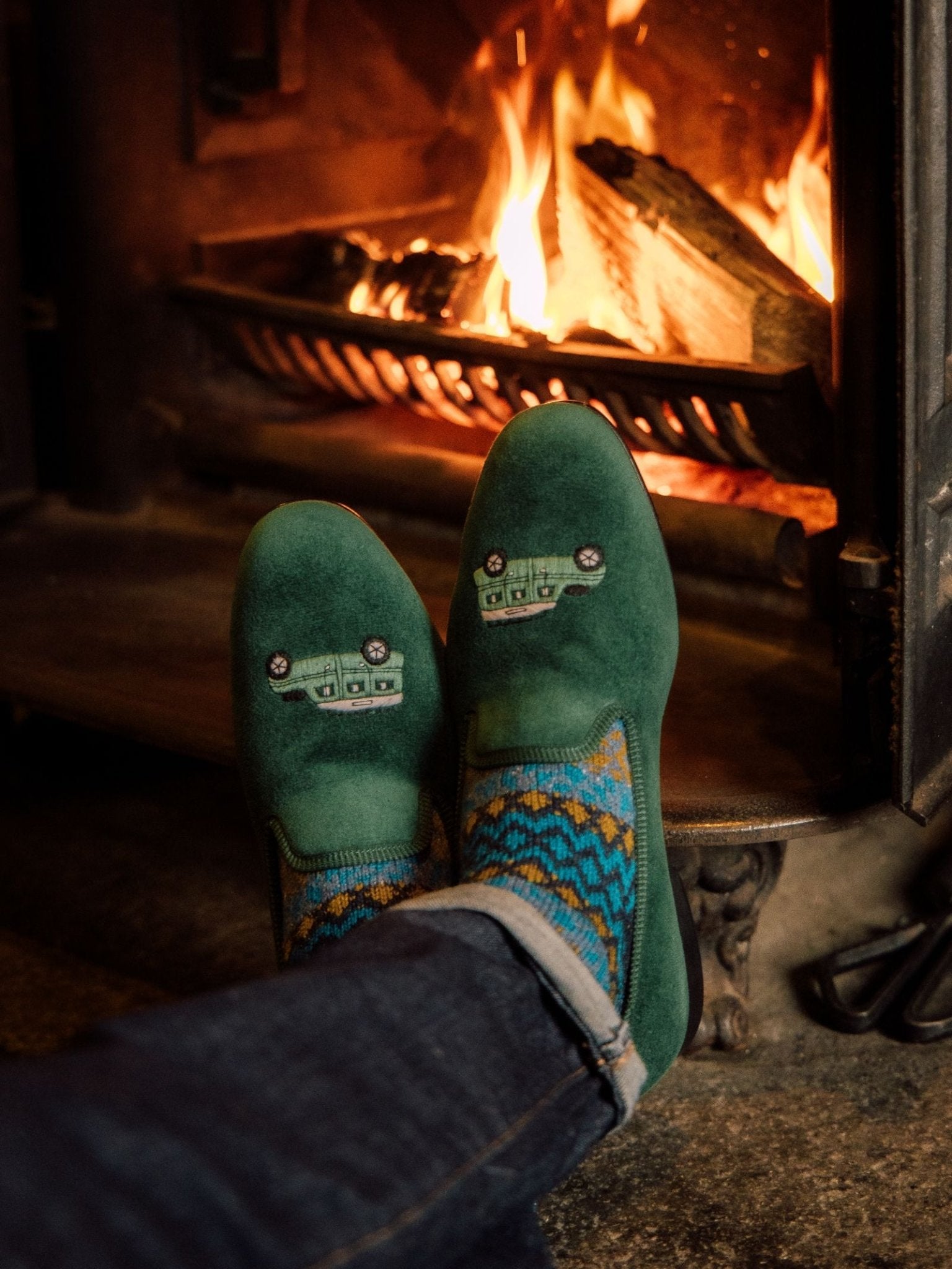 A person in Campbells of Beauly Land Rover Defender Velvet Slippers and colorful socks relaxes by a fireplace with burning logs, creating a warm and cozy atmosphere.