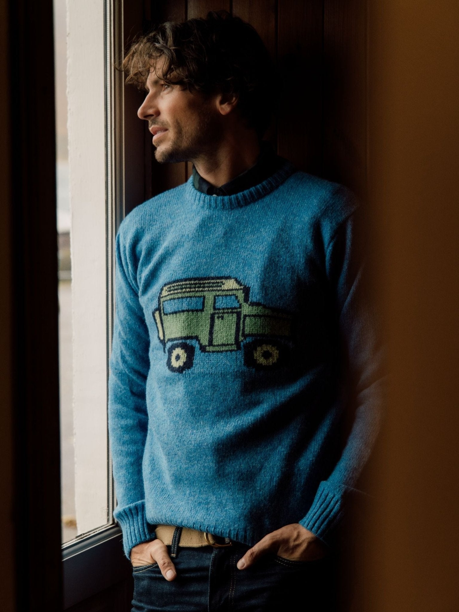 A man stands by a window in a Campbells of Beauly Land Rover Defender Jumper, its classic design evoking the spirit of British motoring adventure as soft light highlights his thoughtful gaze.