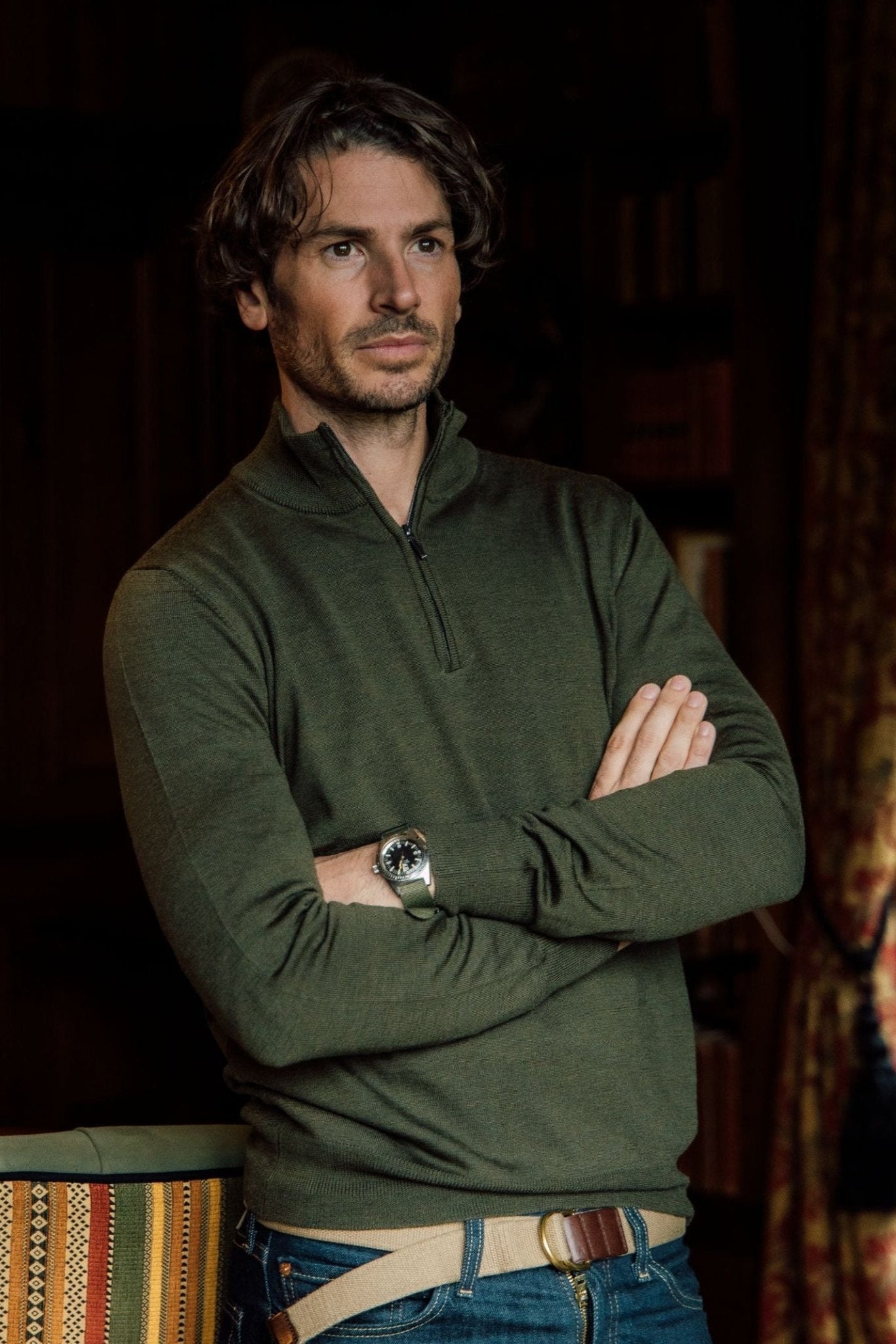 A man with wavy brown hair and light stubble stands indoors, arms crossed, wearing the Campbells of Beauly Merino Quarter Zip Jumper in green—an ideal layering piece—paired with jeans and a watch near a patterned curtain and striped-backed chair.