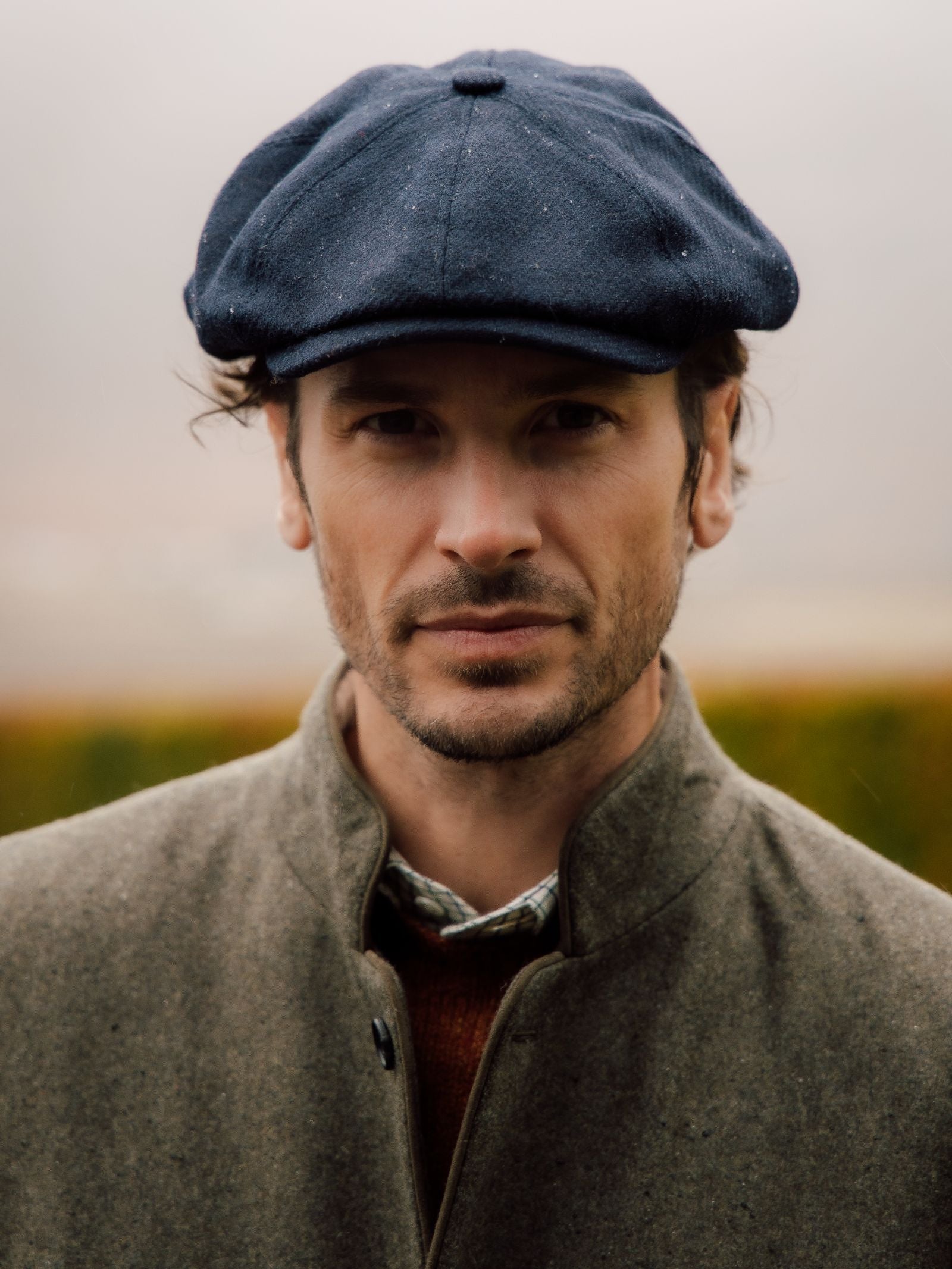 A man with light stubble wears the dark Farrar Cap by Campbells of Beauly and a grey coat, looking directly at the camera with a neutral expression. Blurred greenery and a muted sky evoke British country heritage.