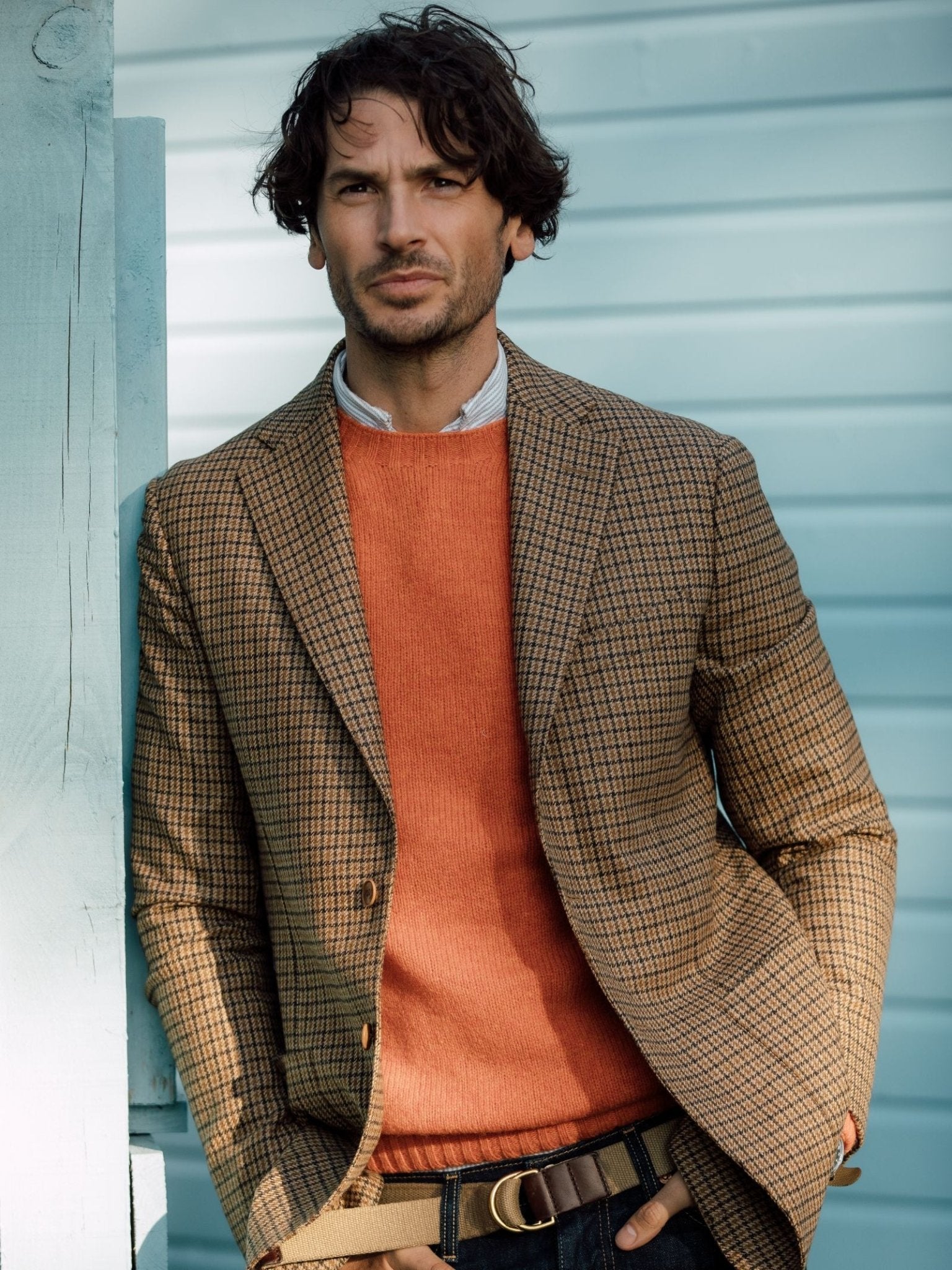 A man with wavy brown hair wears the Campbells of Beauly Gun Club Jacket in brown plaid, an orange sweater, blue collared shirt, and jeans as he stands against a light blue wooden wall, looking at the camera with a neutral expression.