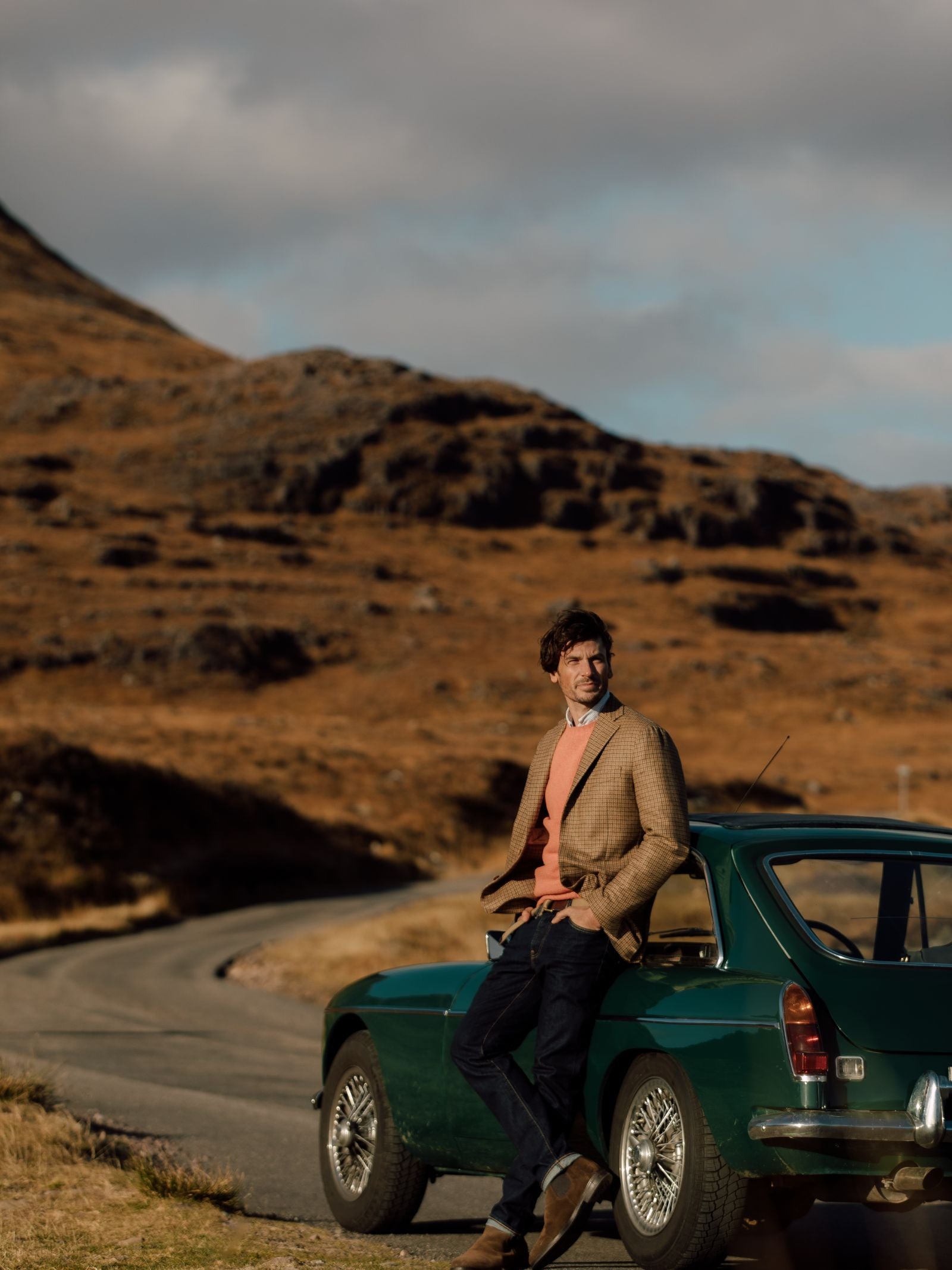 A man in a regular fit Campbells of Beauly Gun Club Jacket and sweater leans against a vintage green car on a winding road, with a sunlit hillside and partly cloudy sky in the background.