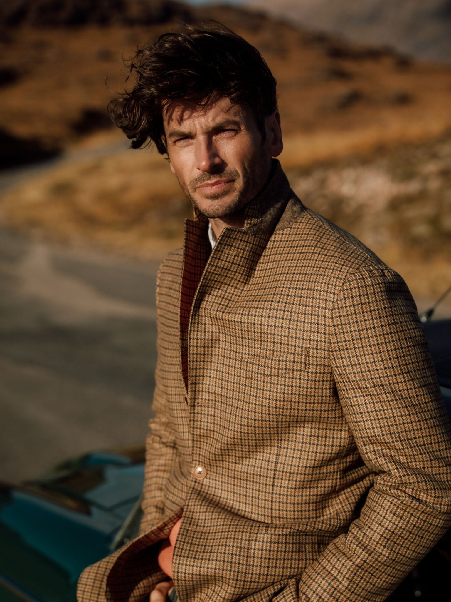 A man with wavy dark hair wears a Campbells of Beauly Gun Club Jacket in a regular fit checked brown pattern, standing outdoors in sunlight against a blurred landscape of hills and a road.
