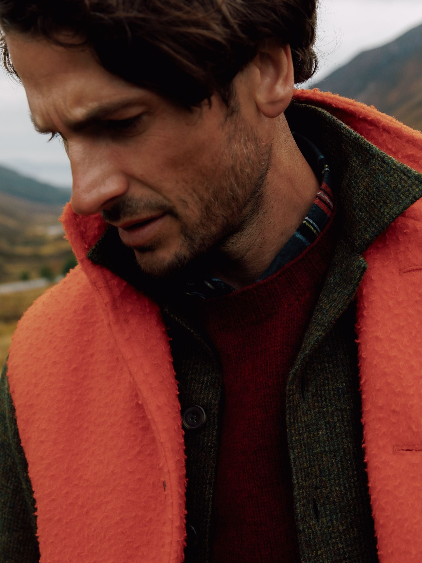 A man with tousled brown hair looks downward, outdoors before a mountain backdrop, wearing a textured orange Casetino Fleece Gilet by Campbells of Beauly over a green jacket and red sweater.