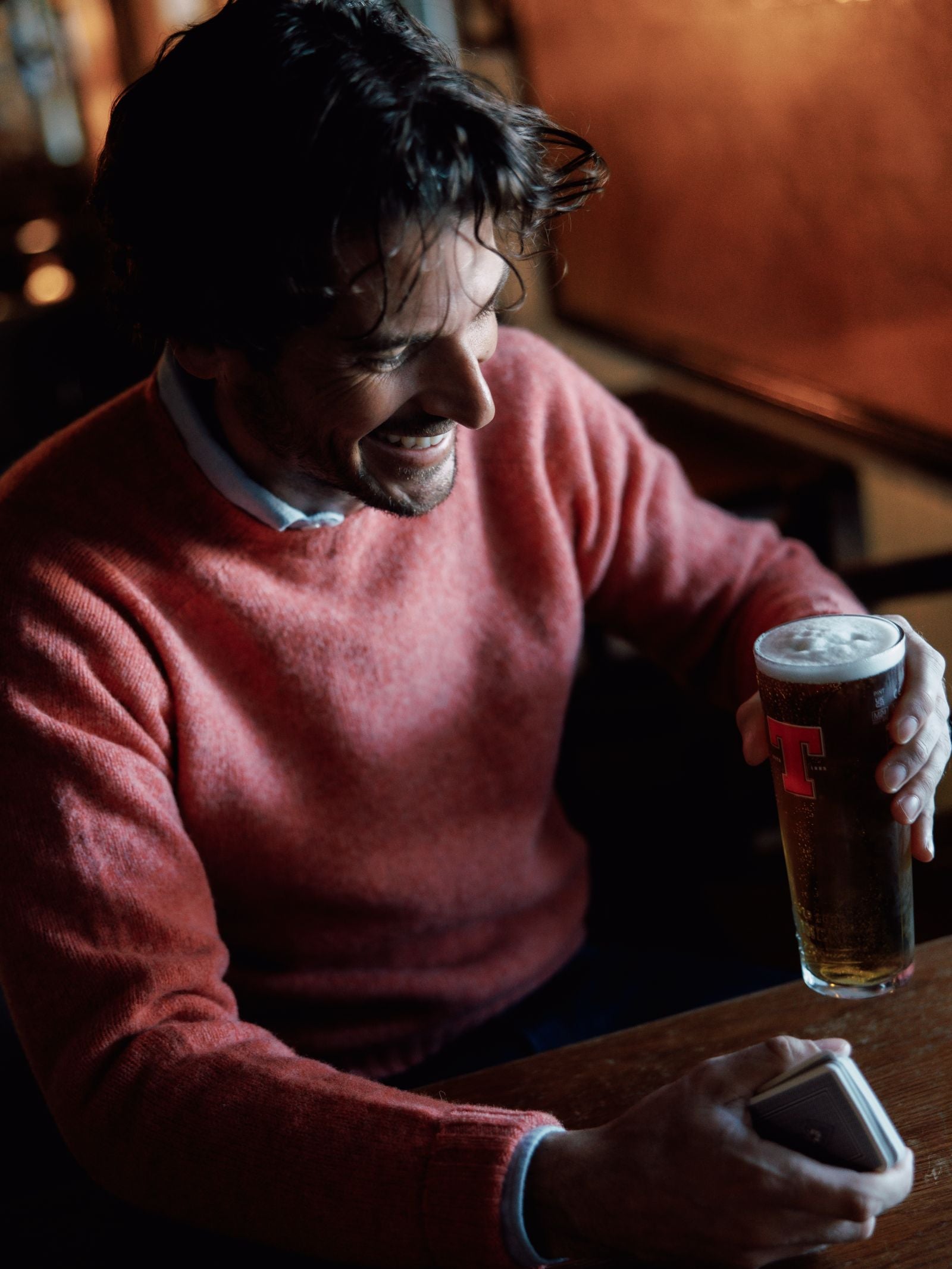 A man wearing a heritage-inspired Shetland Jumper by Campbells of Beauly smiles at his phone, holding a pint with a red T logo, seated at a wooden table in a dimly lit pub.