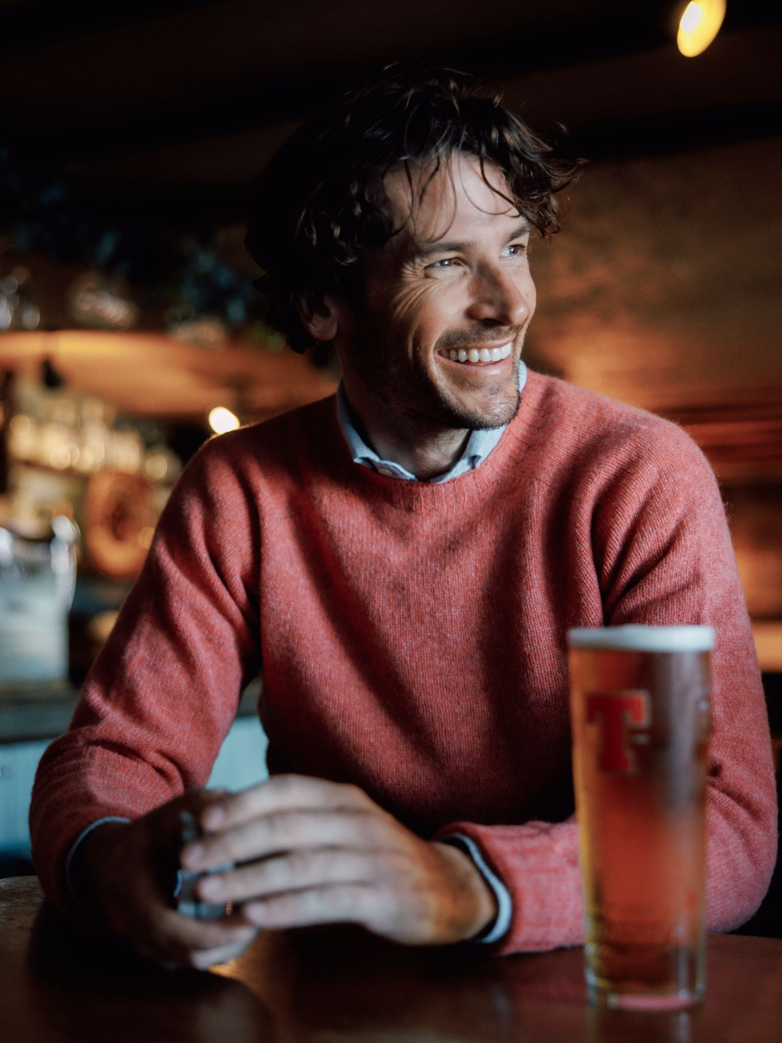 A smiling man with wavy hair wears the Campbells of Beauly Shetland Jumper in a heritage-inspired pink, layered over a collared shirt, as he sits at a warmly lit bar table with a pint of beer in front of him.