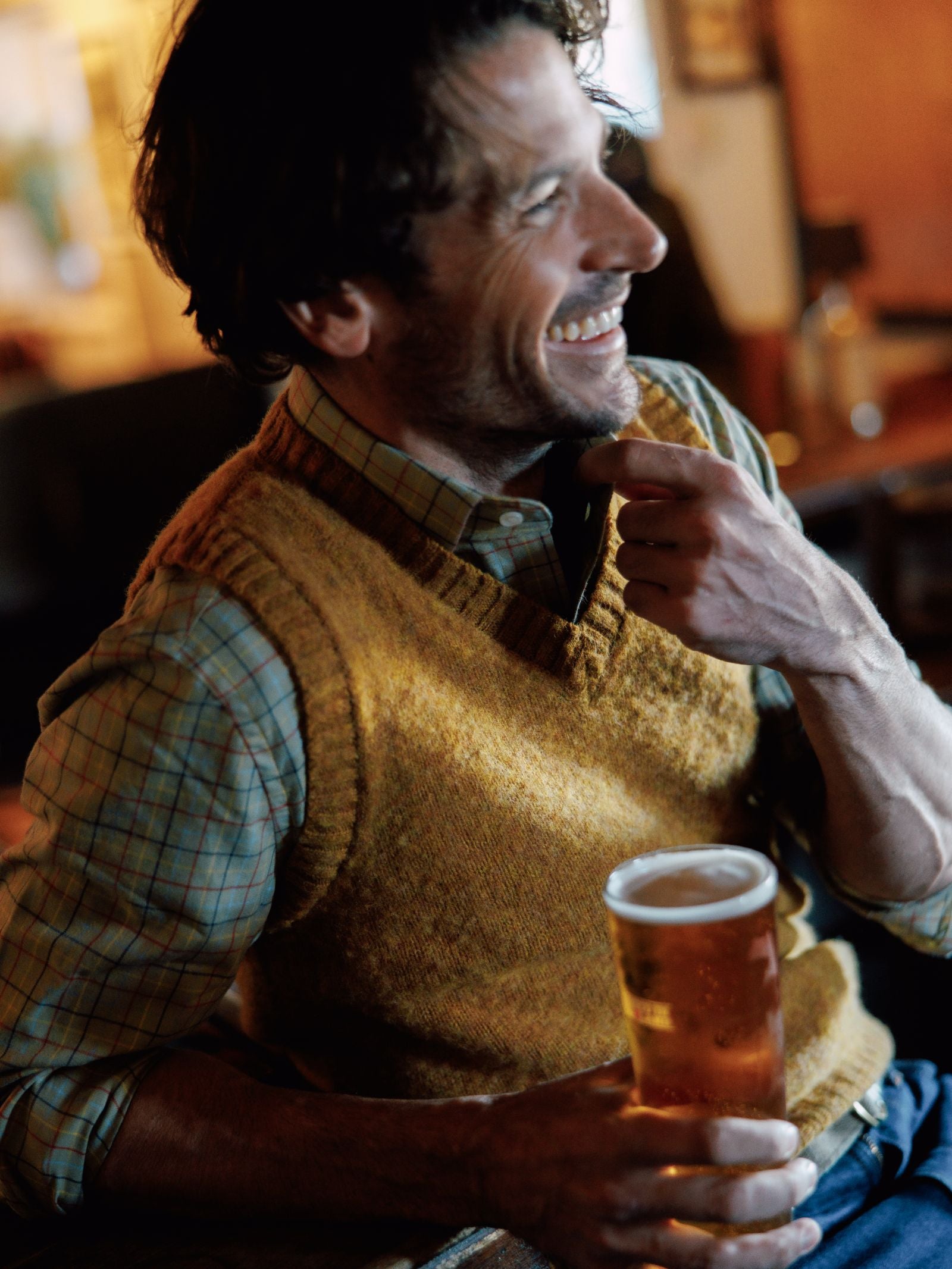 A man in a plaid shirt and a mustard yellow Campbells of Beauly Shetland Slipover smiles indoors, holding a pint of beer. Warm lighting adds to the cozy, relaxed atmosphere.