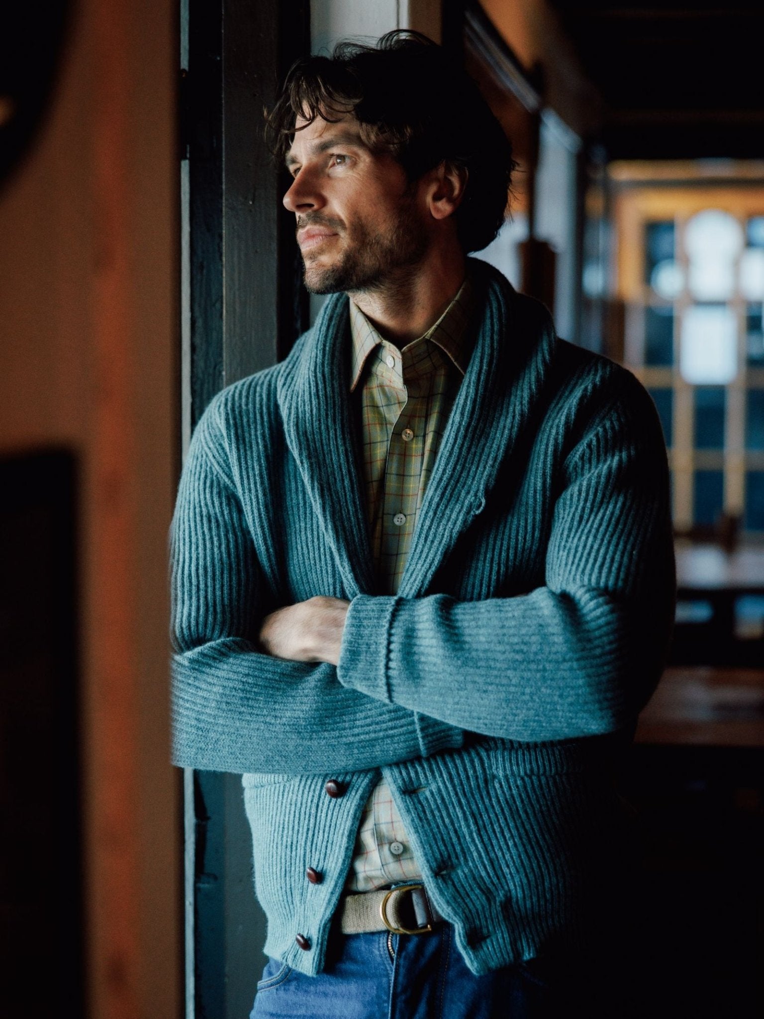 A man with dark hair and a short beard stands indoors, arms crossed, gazing thoughtfully out the window in a blue Shawl Collar Cardigan by Campbells of Beauly—a classic winter essential.