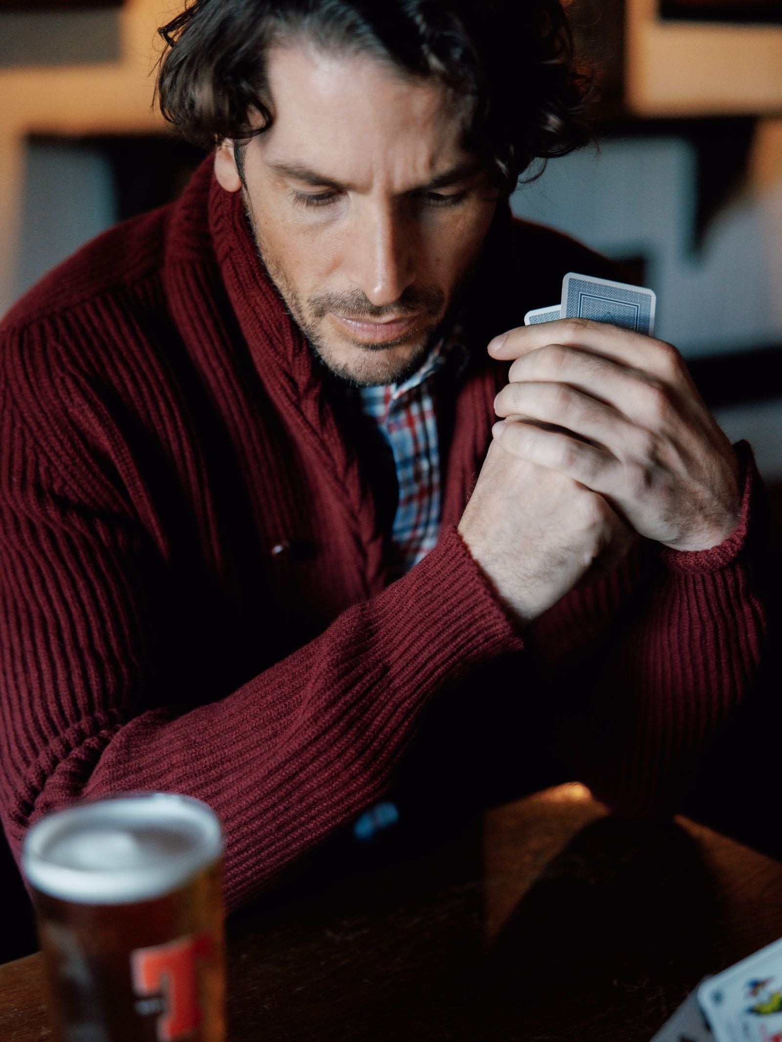 A man in a Campbells of Beauly Shawl Collar Cardigan, a winter wardrobe essential, sits at a wooden table with a deck of cards and a glass of beer, gazing down thoughtfully.