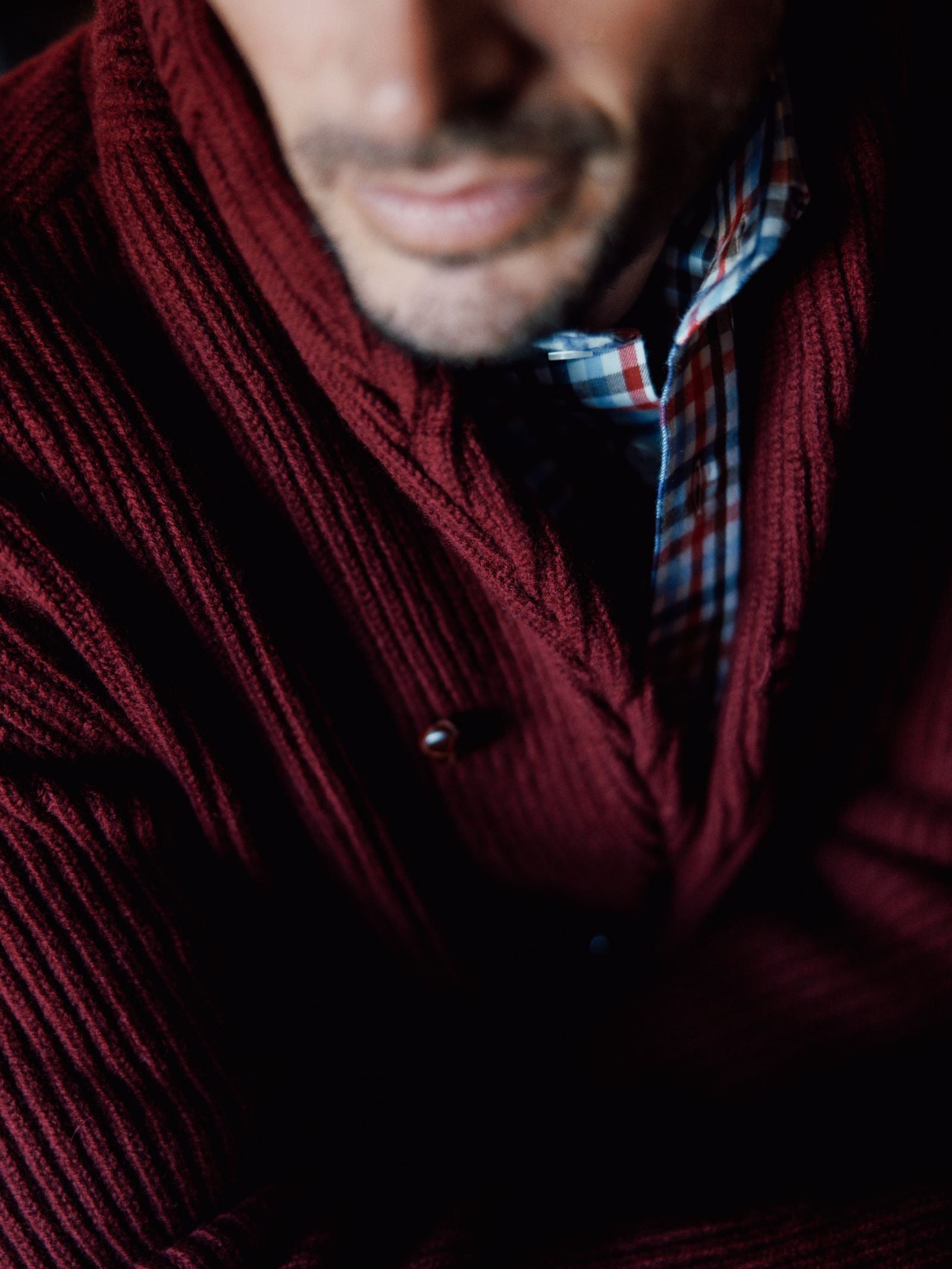 A close-up shows a man wearing the Campbells of Beauly Shawl Collar Cardigan in burgundy layered over a blue, red, and white plaid shirt. His lower face, mouth, and beard are visible; the rest is softly out of focus.