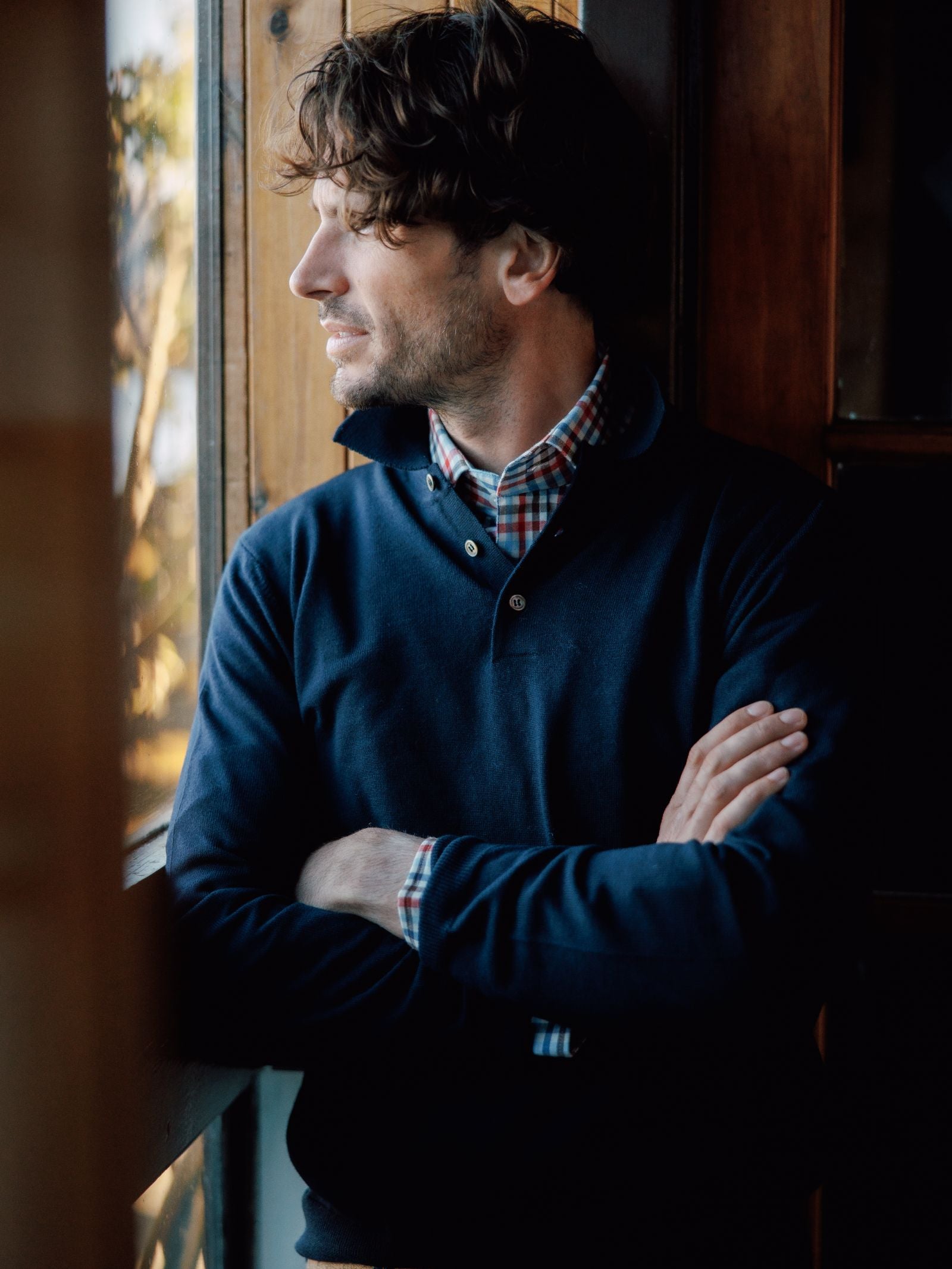 A man with wavy brown hair and a short beard stands indoors by a wooden window, arms crossed, gazing thoughtfully outside while wearing the Campbells of Beauly Merino Shirt Jumper over a plaid shirt.