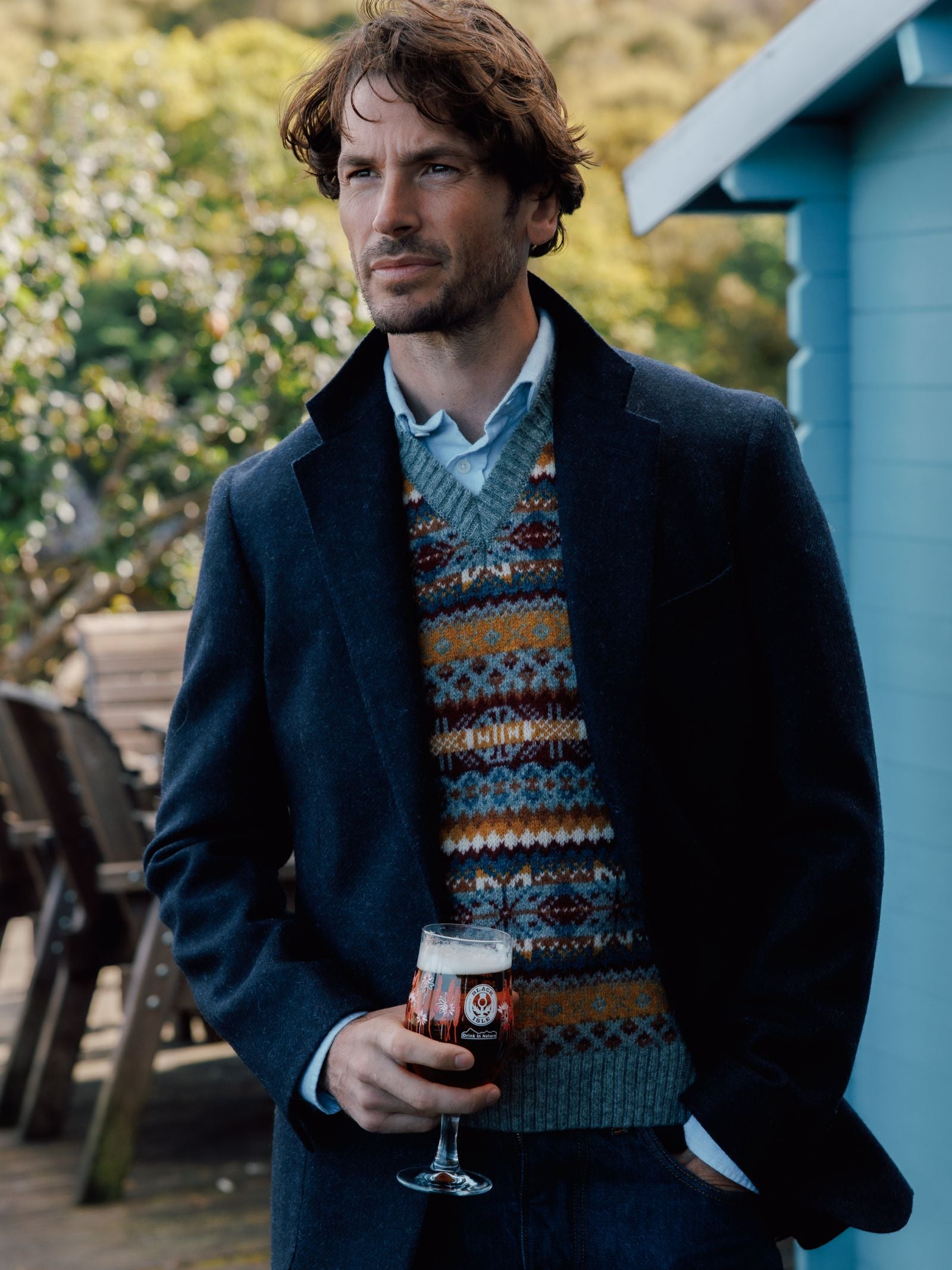 A man with wavy brown hair wears a Campbells of Beauly Navy Twill House Jacket over a patterned sweater and blue shirt, standing outside by a blue building, holding dark beer, with trees and wooden chairs in the background.
