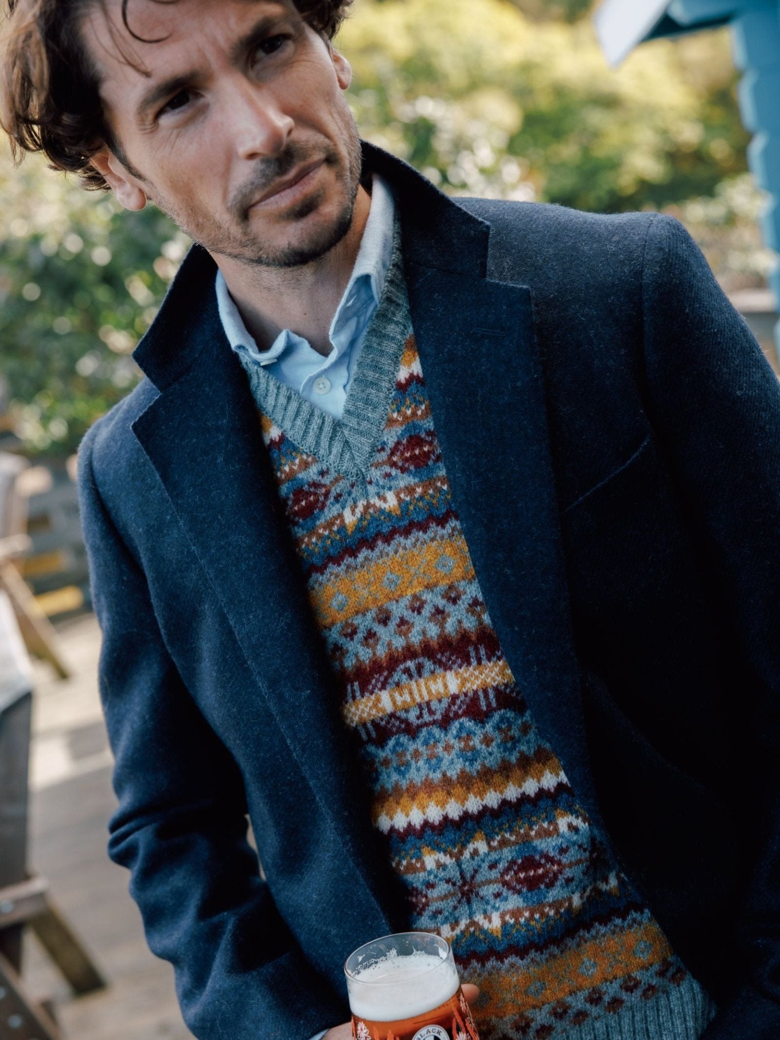 A man with wavy brown hair wears the Campbells of Beauly Navy Twill House Jacket over a colorful Shetland wool sweater and light blue shirt, standing outdoors with a pint of beer. Trees and a blue building blur in the background.