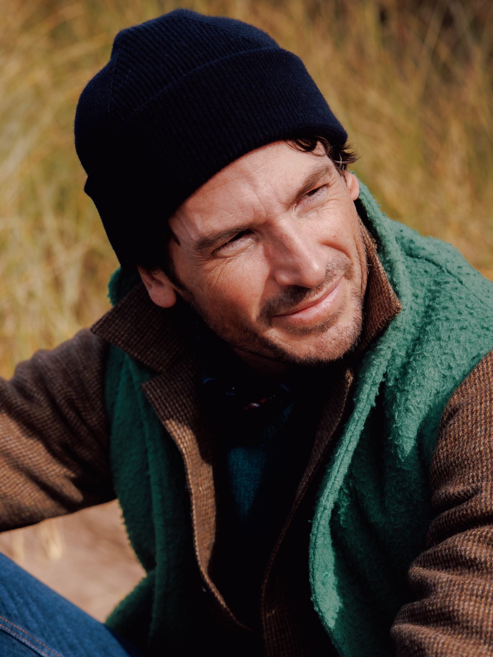 A man wearing a black knit beanie, brown jacket, and a green Campbells of Beauly Casetino Fleece Gilet sits outdoors, smiling slightly with dry grass blurred in the background.
