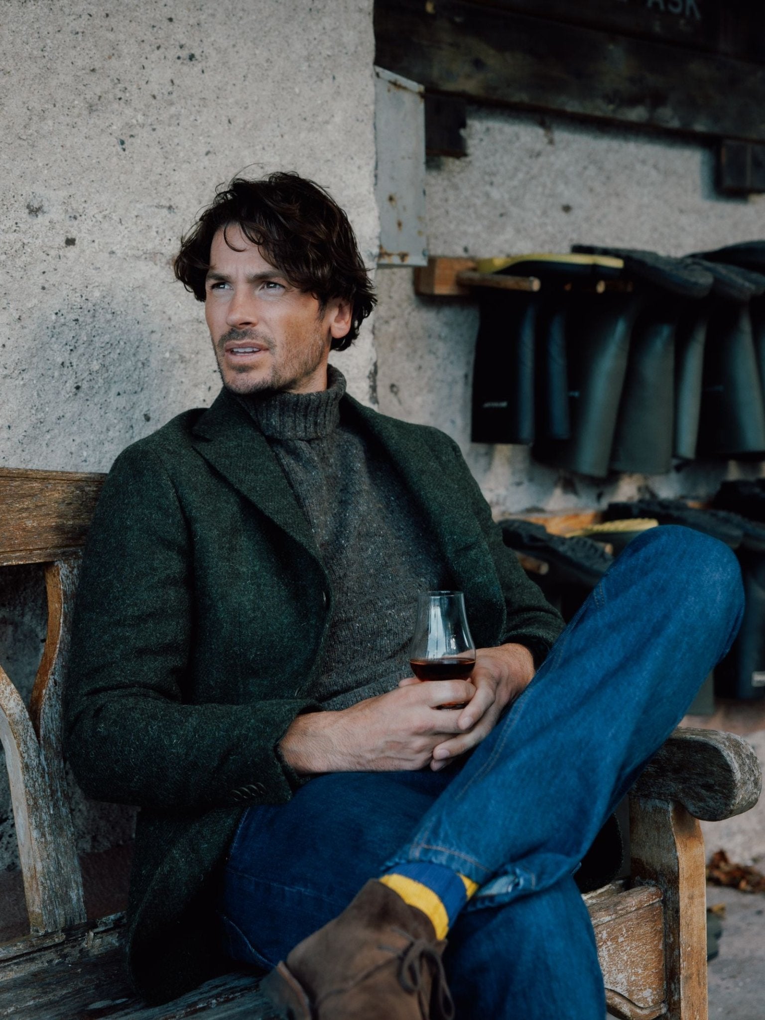 A man with wavy brown hair sits on a wooden bench, holding a glass. He wears a Campbells of Beauly Harris Tweed Jacket over a gray sweater, blue jeans, and brown shoes, with rubber boots neatly arranged on shelves behind him.