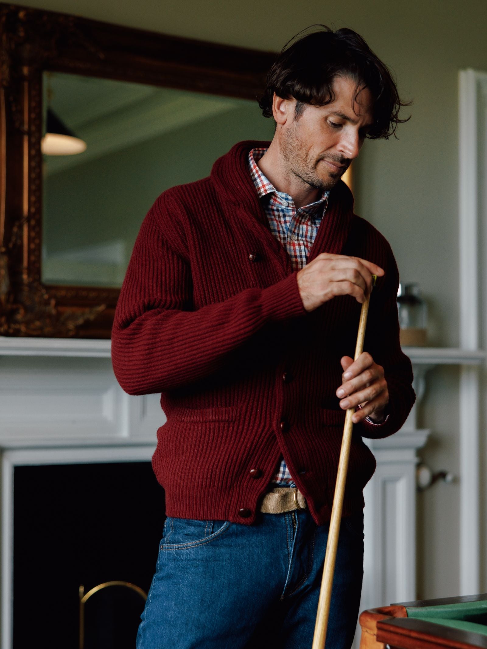 A man with wavy dark hair wears the Campbells of Beauly Shawl Collar Cardigan over a plaid shirt and jeans, holding a pool cue. He stands by a fireplace and ornate mirror, capturing a cozy winter look.