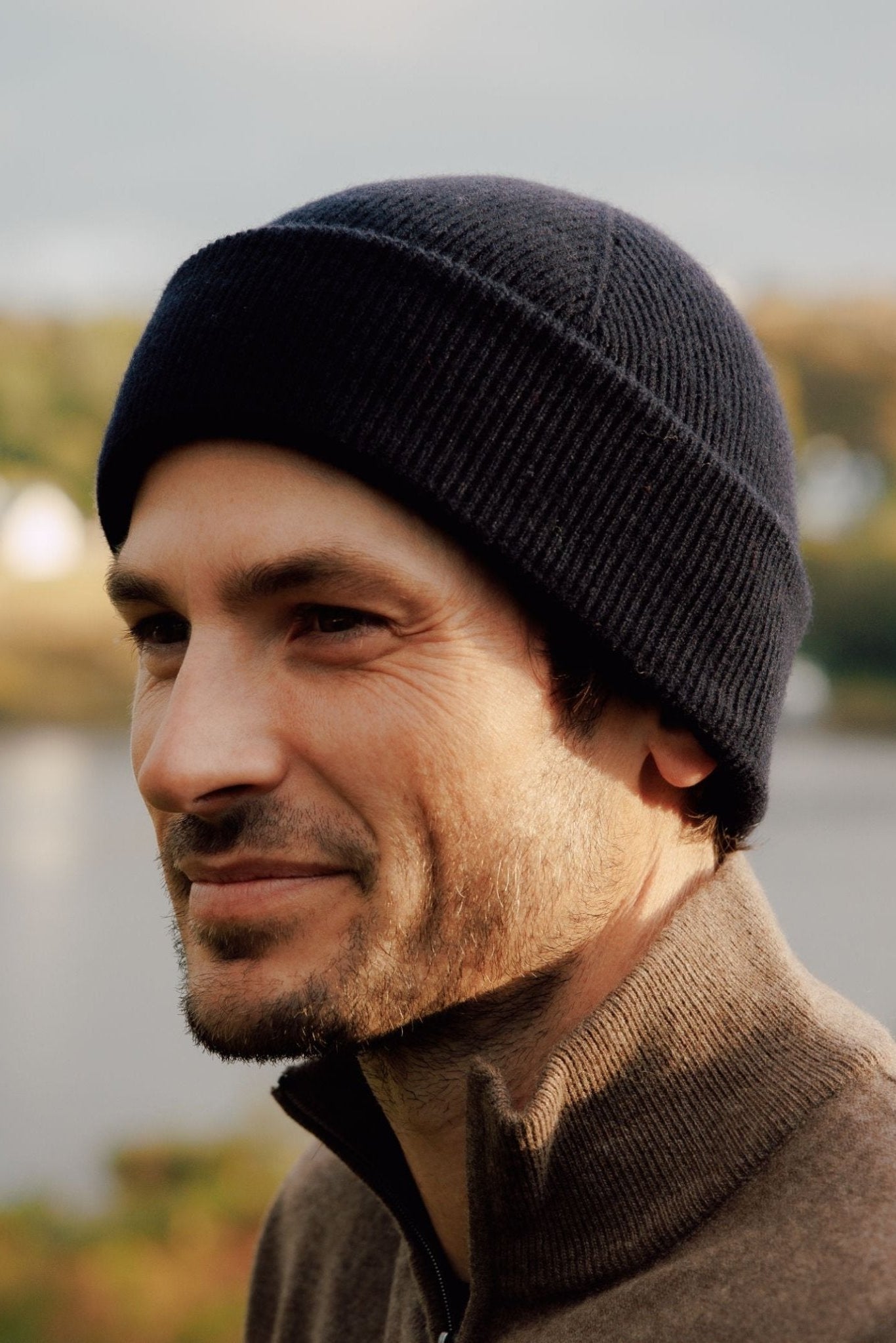 A man with light stubble smiles softly while looking to the side outdoors in the Scottish Borders, wearing a dark Campbells of Beauly Watchman Hat and a brown zip-up sweater, against a blurred natural background.