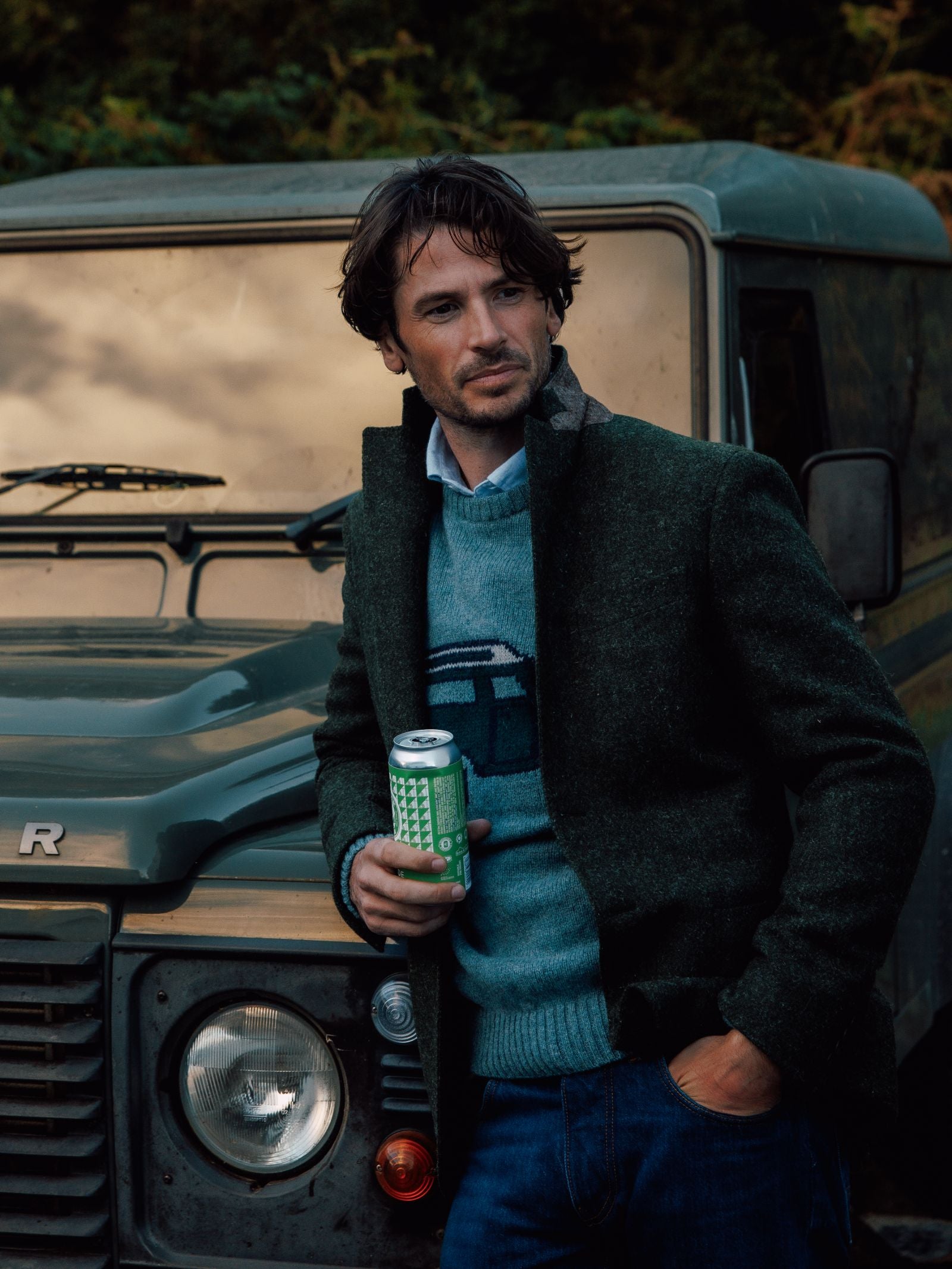 A man in a Land Rover Defender Jumper by Campbells of Beauly stands before a Defender, exuding classic British motoring style as he holds a can and looks to the side with one hand in his pocket.