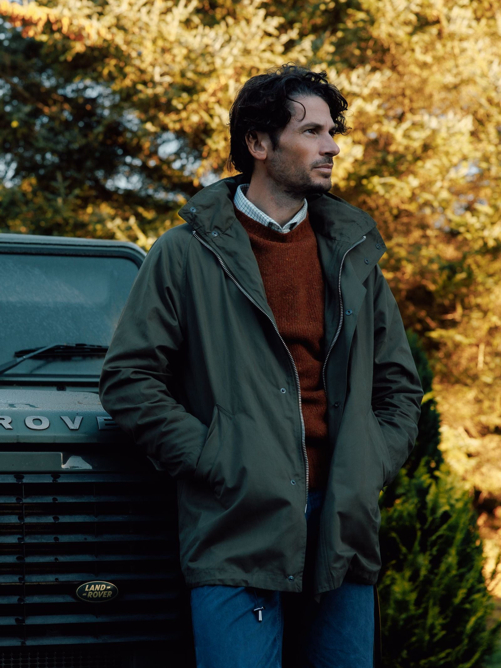 A man with wavy dark hair stands beside a green Land Rover, wearing a Campbell’s of Beauly Mont Coat over a rust sweater and blue jeans. Behind him, autumn trees glow in golden sunlight, creating a warm, natural backdrop.