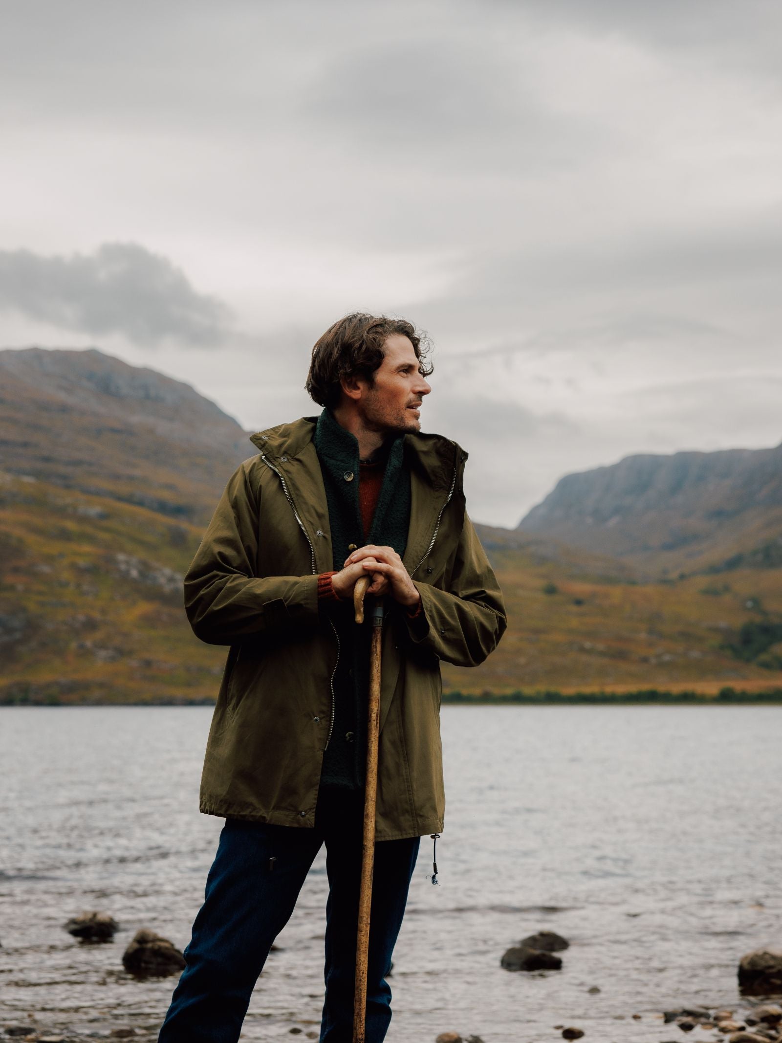 A man in a Campbells of Beauly Mont Coat stands by a lakeside, holding a wooden stick and gazing to the side. Behind him are mountains and clouds, with greenery and rocks lining the rugged shore.