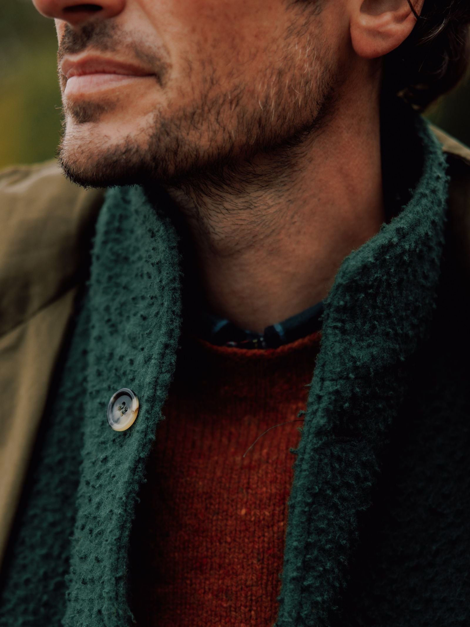 A close-up of a man’s lower face and neck, showing stubble. He wears a green Casetino Fleece Gilet by Campbells of Beauly over a rust sweater, with a patterned shirt visible at the collar.