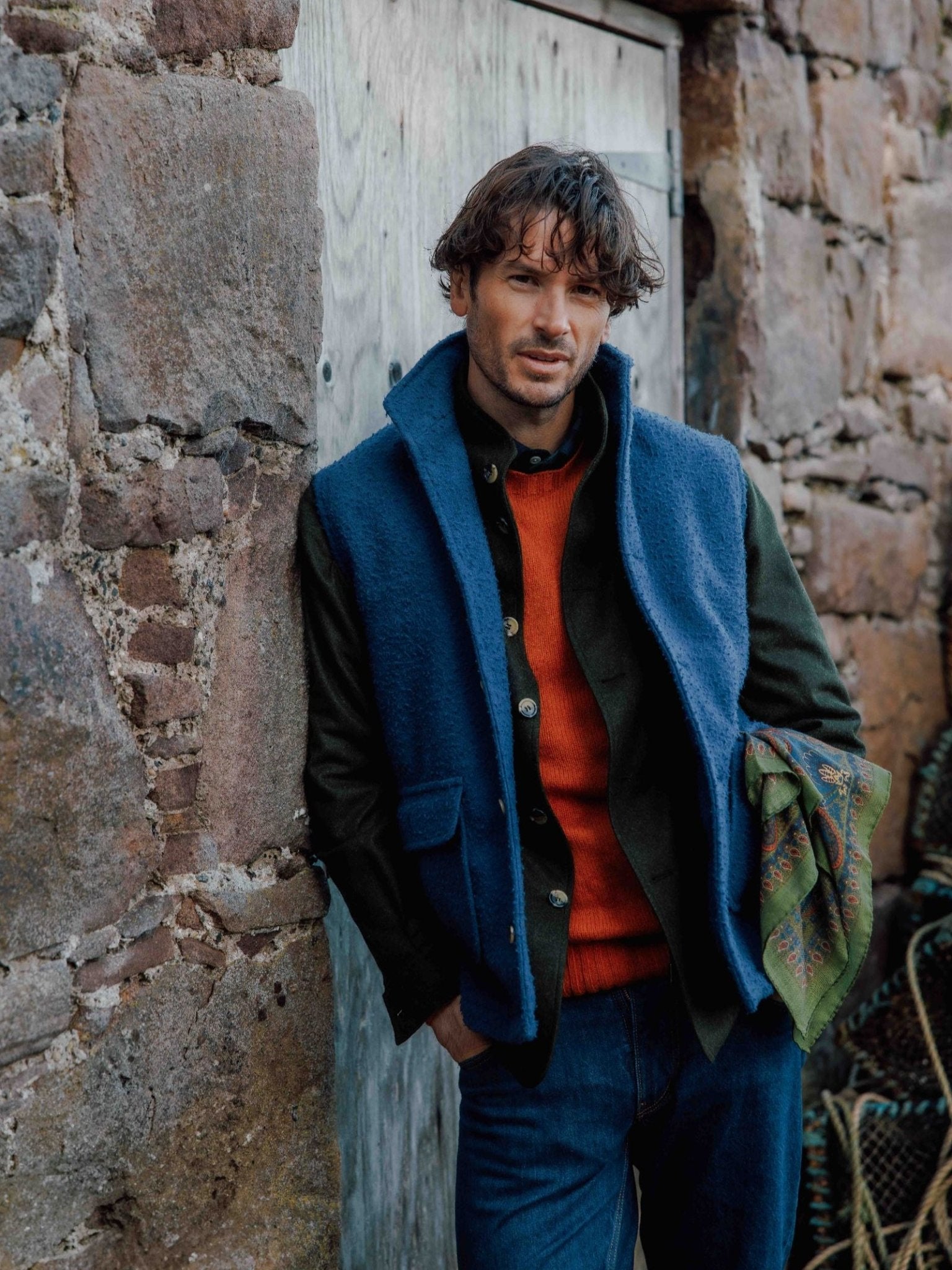 A man with wavy hair leans against a stone wall and wooden door, wearing the Campbell's of Beauly Casentino Fleece Gilet over a red sweater and dark shirt, paired with blue jeans and holding a patterned green scarf.