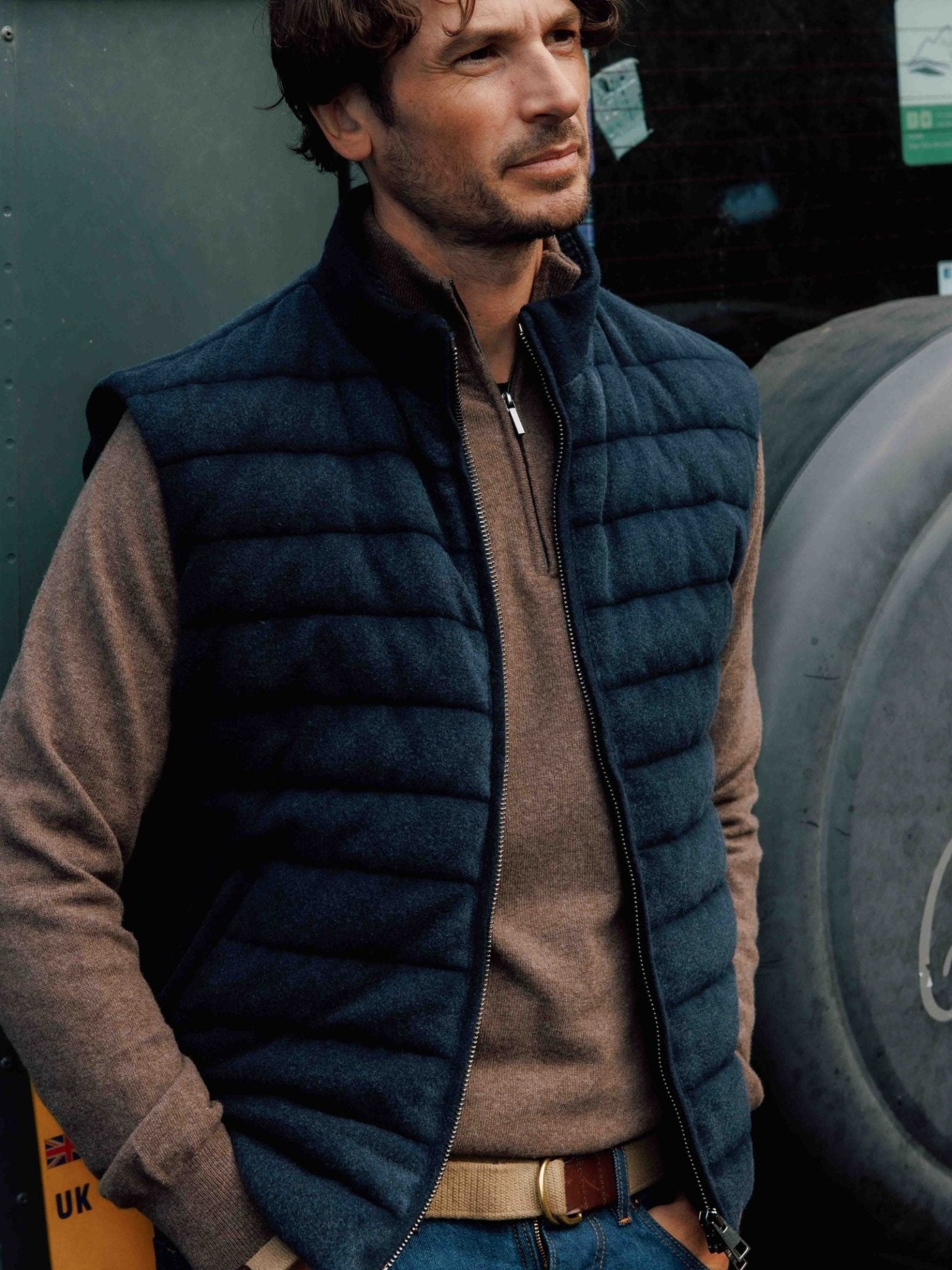 A man with wavy brown hair wears the Campbells of Beauly Quilted Navy Zip Gilet over a brown merino-cashmere blend zip sweater and jeans, standing outdoors with hands in pockets, showcasing effortless layering.