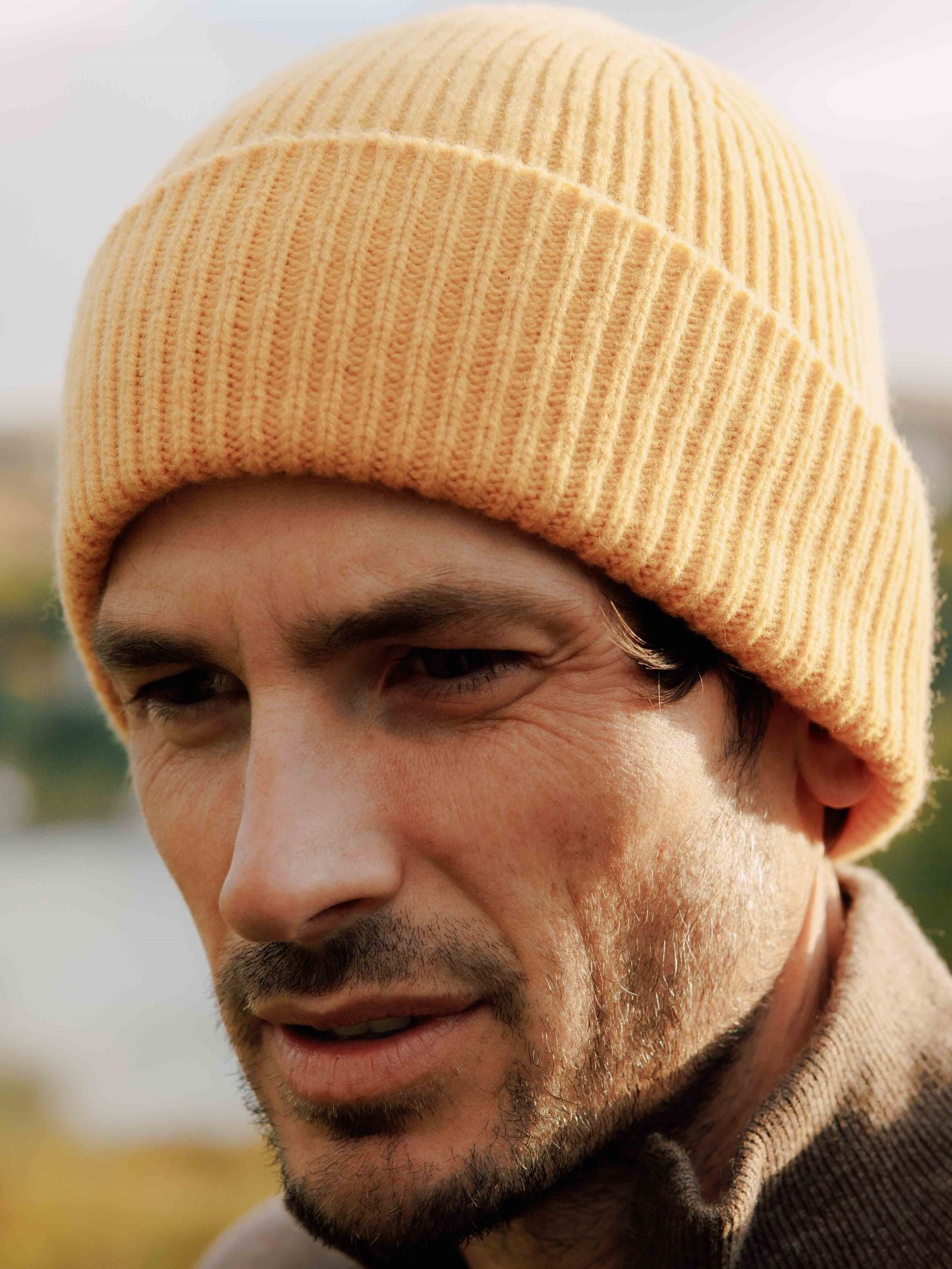 A man with stubble wears the Campbells of Beauly Lambswool Rib Hat in mustard yellow and a brown jacket, gazing downward thoughtfully. The softly blurred background suggests the Scottish Borders, evoking cozy winter style.