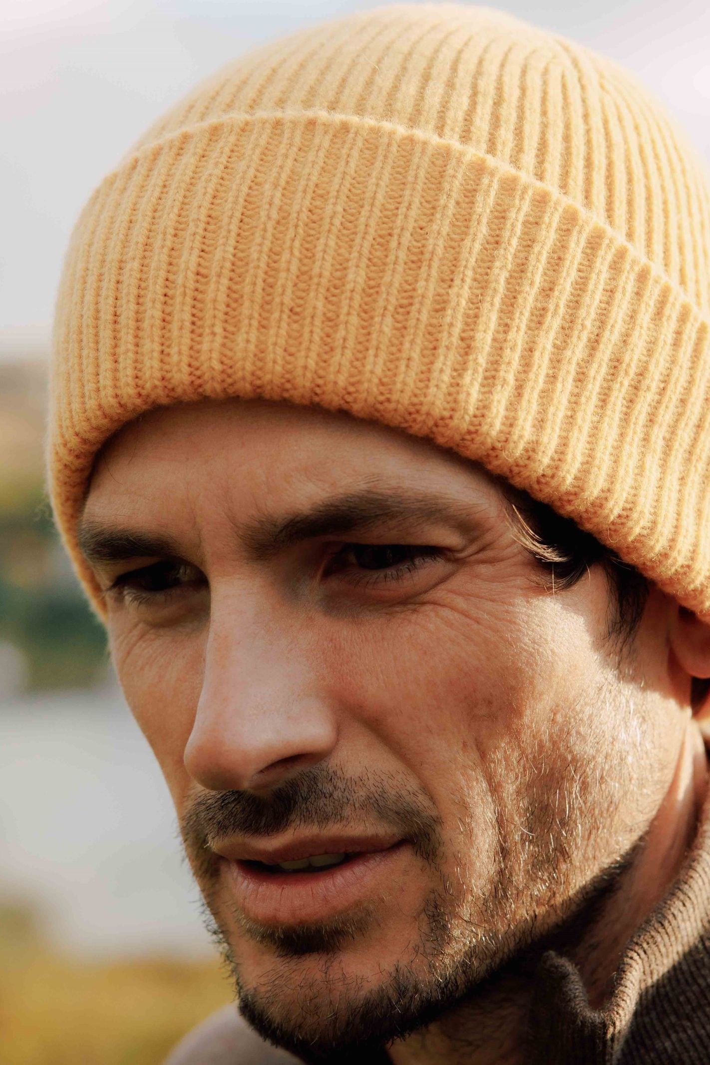 A man with stubble wears the Campbells of Beauly Lambswool Rib Hat in mustard yellow and a brown jacket, gazing downward thoughtfully. The softly blurred background suggests the Scottish Borders, evoking cozy winter style.