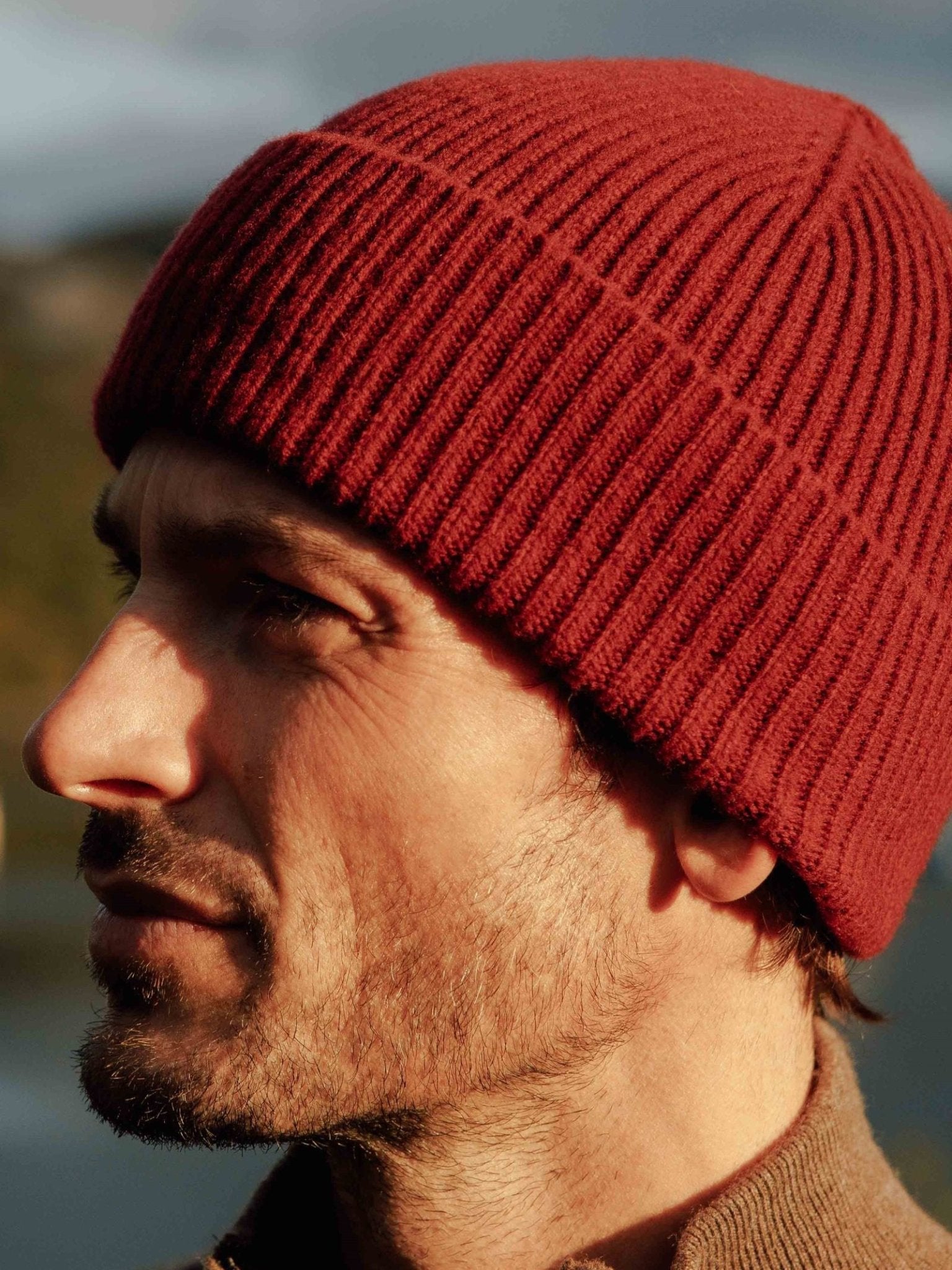 A man with stubble wears the Campbells of Beauly Lambswool Rib Hat—a cozy red hat perfect for winter—while gazing into the distance outdoors in the Scottish Borders, set against a softly blurred background.