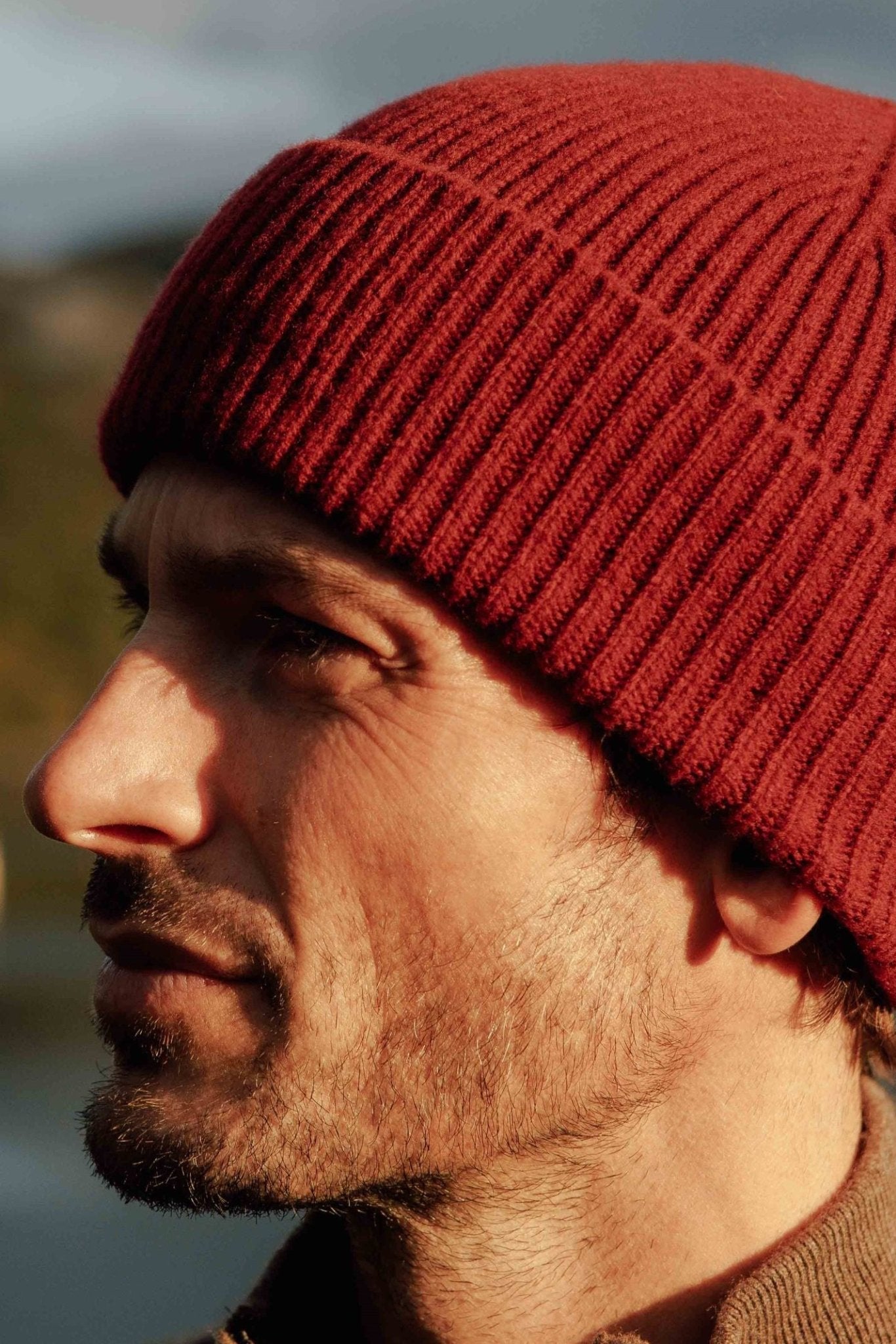 A man with stubble wears the Campbells of Beauly Lambswool Rib Hat—a cozy red hat perfect for winter—while gazing into the distance outdoors in the Scottish Borders, set against a softly blurred background.