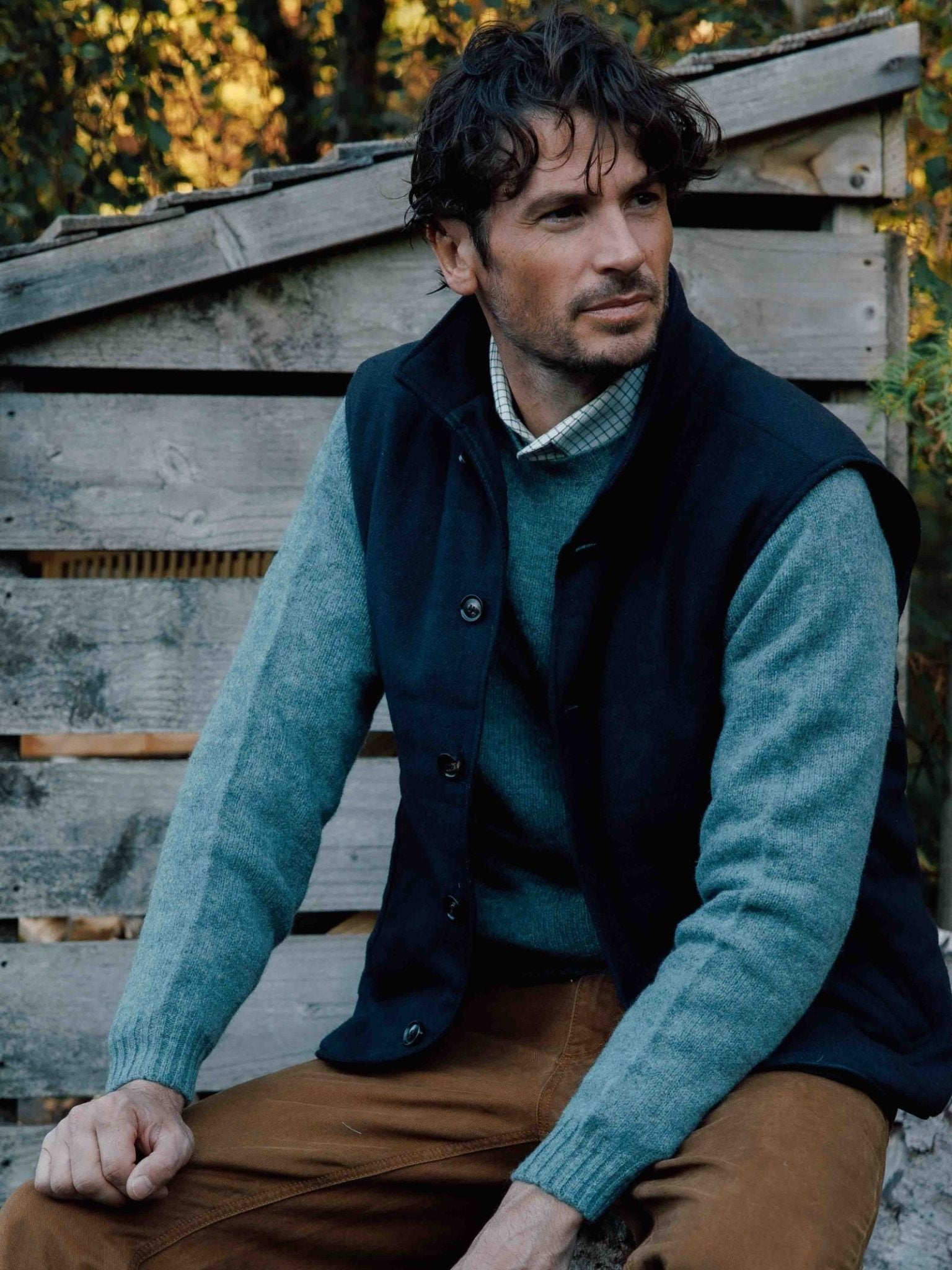 A man with wavy dark hair sits outdoors by a rustic wooden structure, wearing a blue sweater, brown pants, and the Campbells of Beauly Quilted Tweed Buttoned Gilet as a stylish layering piece, gazing thoughtfully to the side.