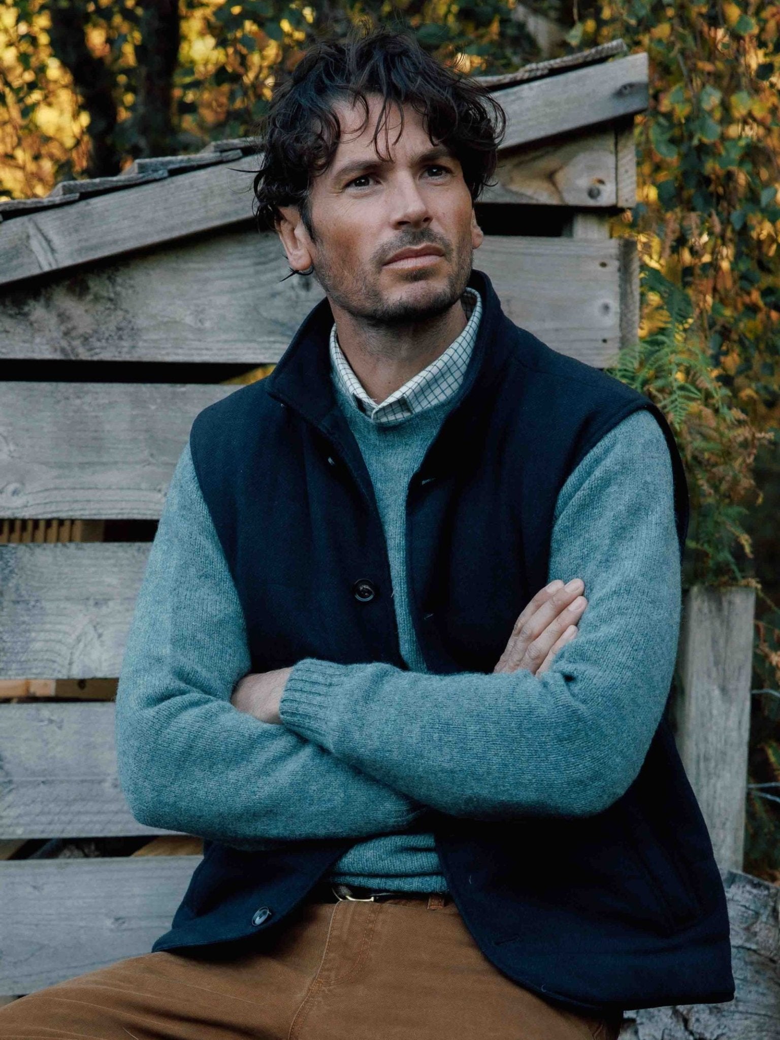 A man with wavy dark hair sits outdoors in front of a rustic wooden structure and greenery, wearing a blue sweater, brown pants, and the Campbells of Beauly Quilted Tweed Buttoned Gilet—a classic layering piece.