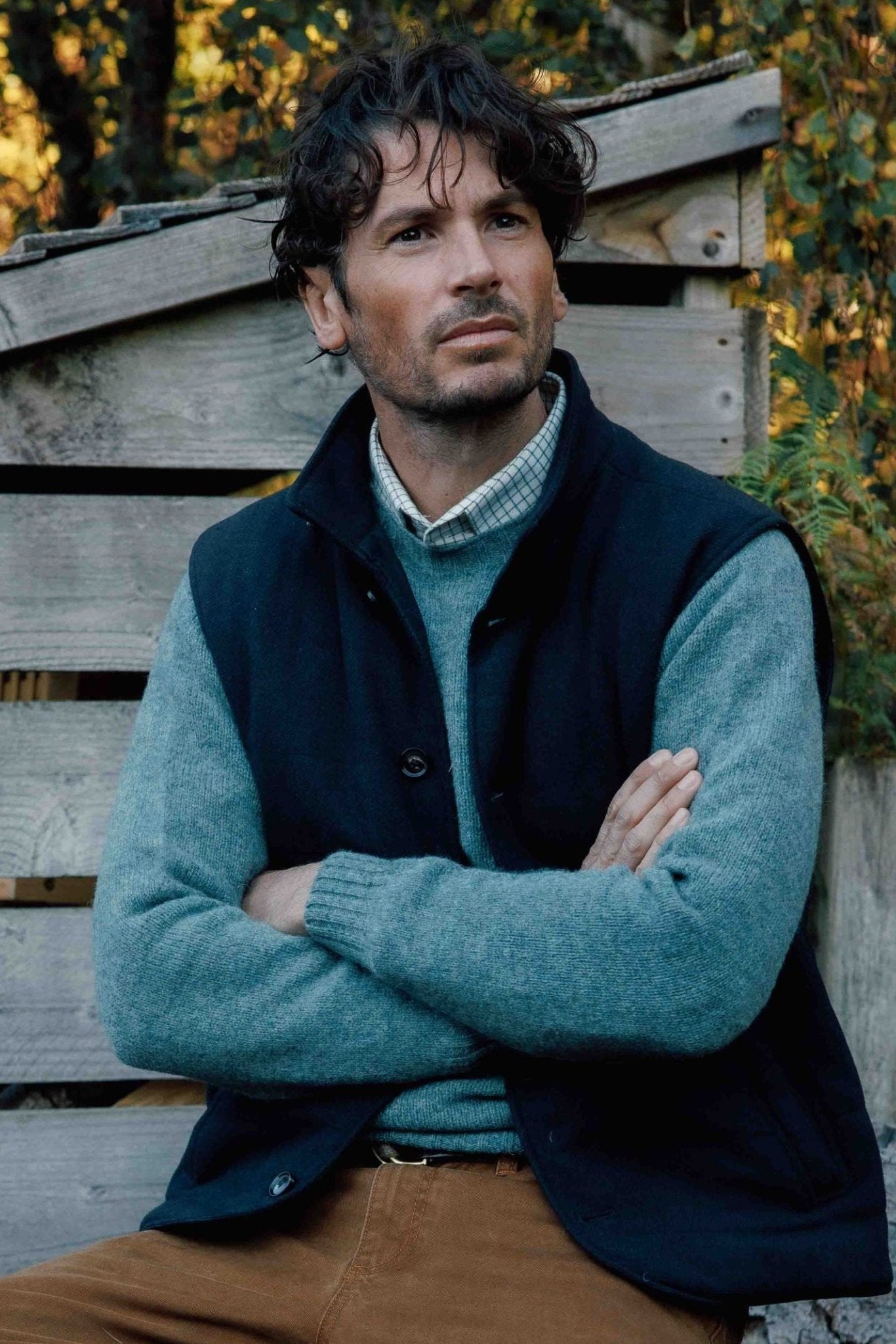 A man with wavy dark hair sits outdoors in front of a rustic wooden structure and greenery, wearing a blue sweater, brown pants, and the Campbells of Beauly Quilted Tweed Buttoned Gilet—a classic layering piece.