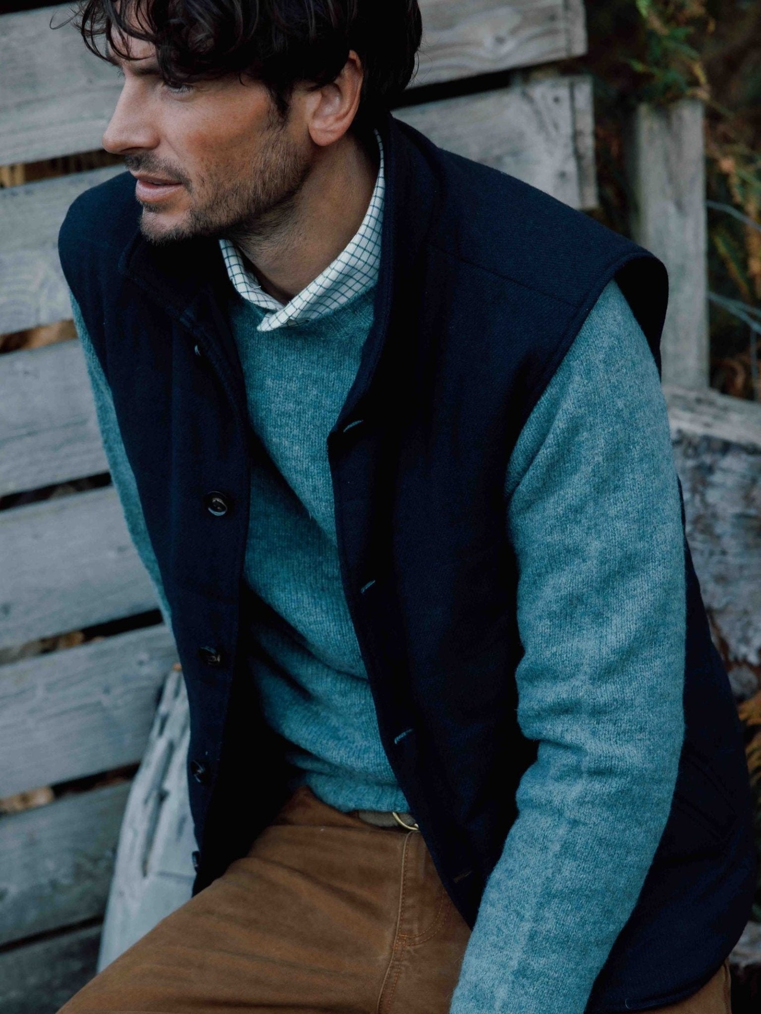 A man with dark hair and light stubble relaxes outdoors on a wooden bench, wearing Campbell’s of Beauly’s Quilted Tweed Buttoned Gilet layered over a blue sweater and checked shirt, paired with brown pants. He gazes to the side calmly.