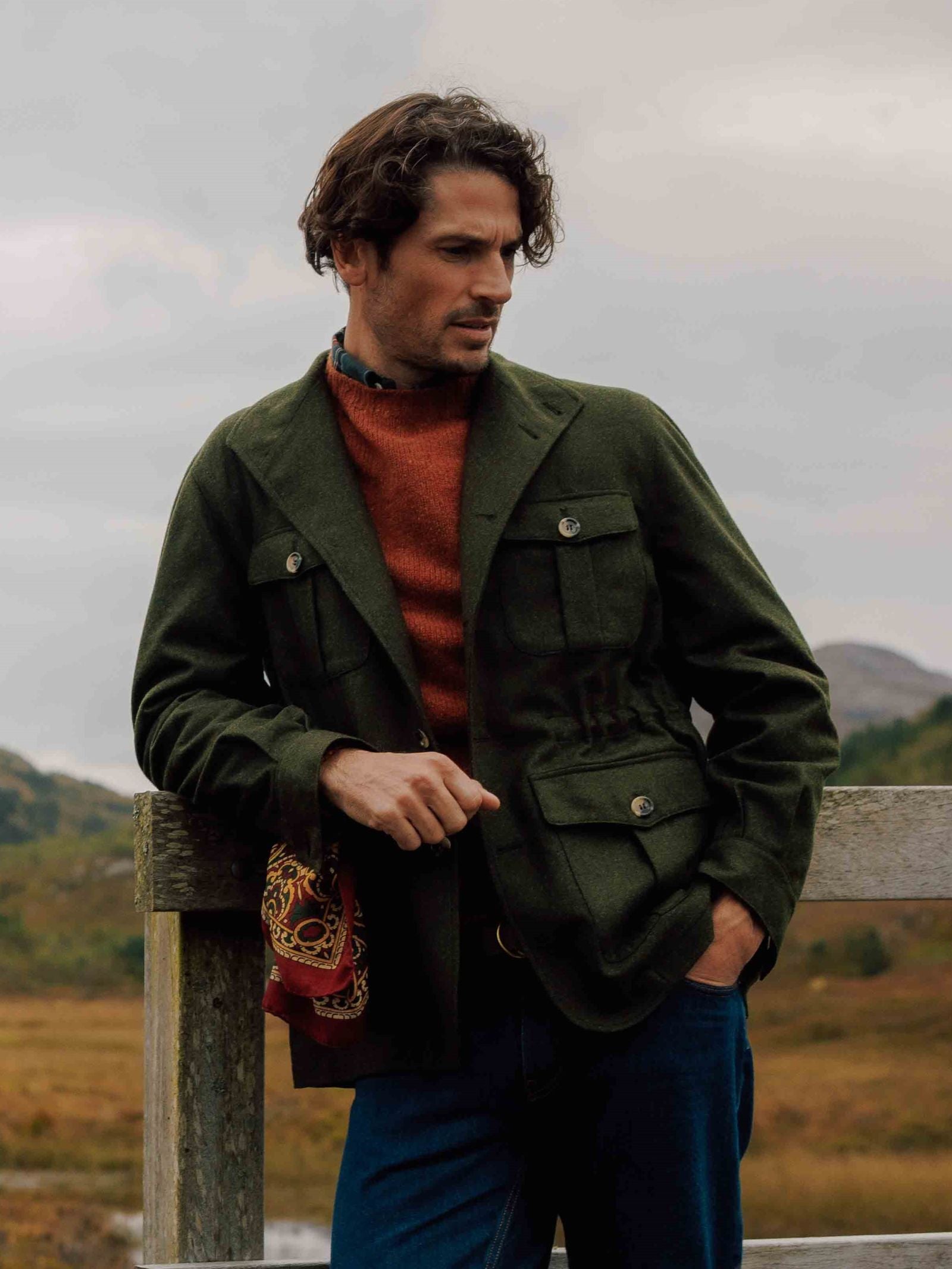 A man with wavy brown hair wears the Campbells of Beauly Field Teba Jacket in green over an orange sweater, leaning on a wooden fence with scarf in hand, backed by a scenic cloudy landscape and classic wool Austrian loden.